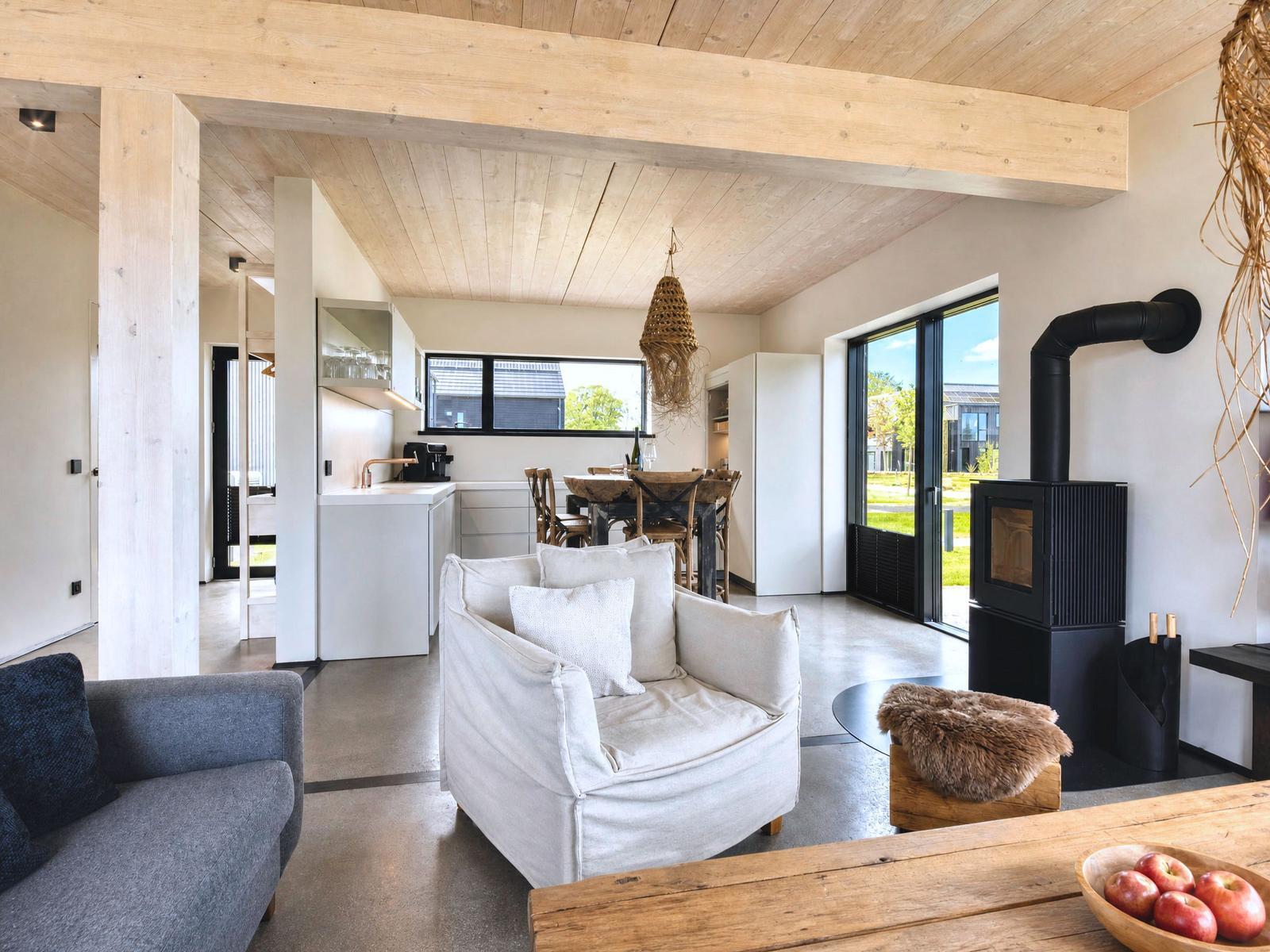 Living area with sofa, fireplace, and kitchen. Wooden ceiling and large windows.