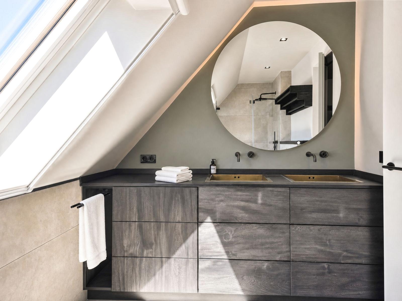 Bathroom with sloped ceiling, round mirror, and sinks.
