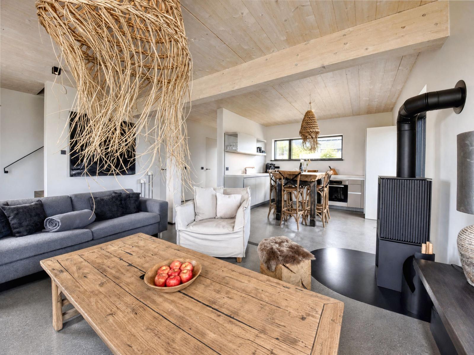Living area with sofa, armchair, and wooden table. Hanging lamp, fireplace, and kitchen visible.