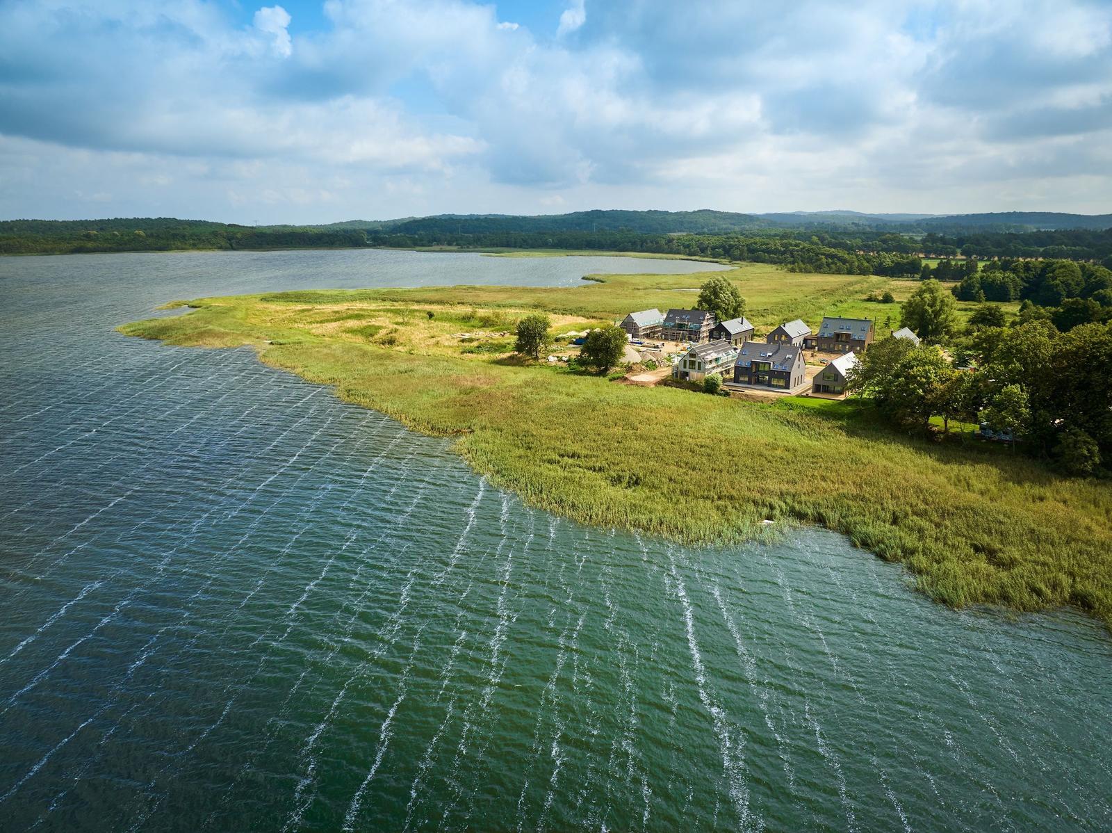 Houses by a lake with green areas and forest in the background.
