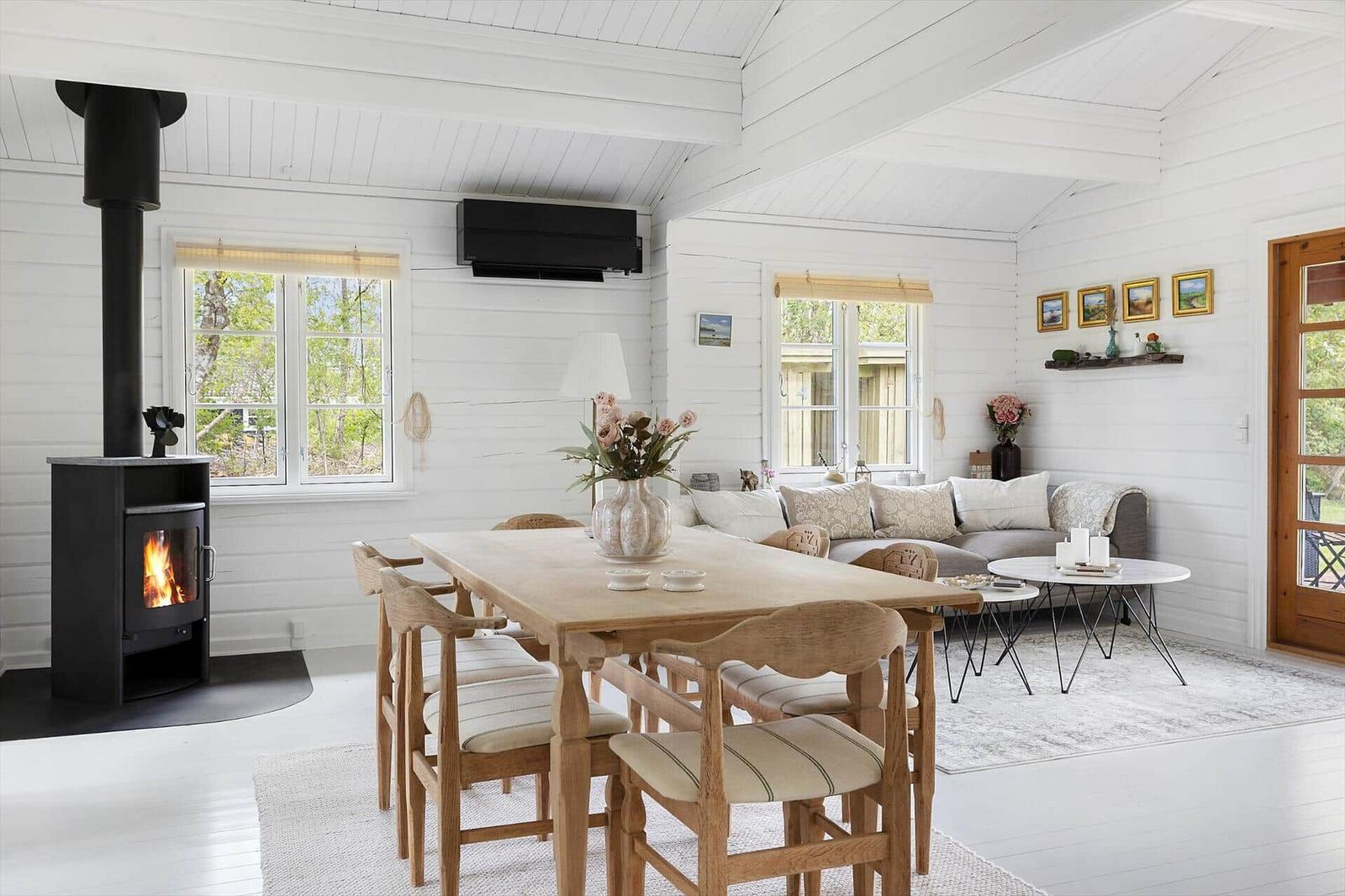 A room with dining table, sofa, and wood stove. Windows and white walls.