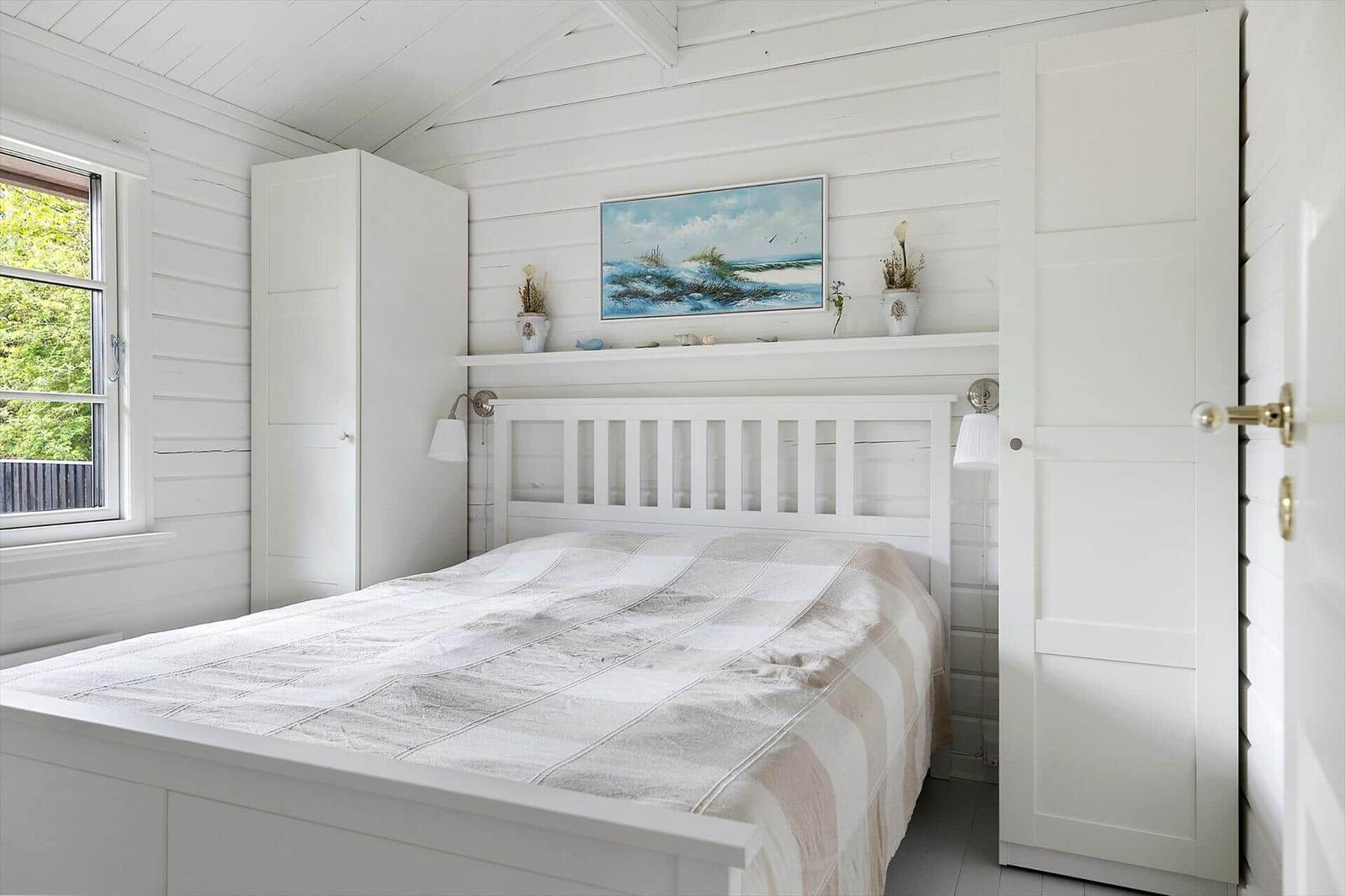 Bedroom with white bed, wall painting, and window. Wooden floor and white walls.