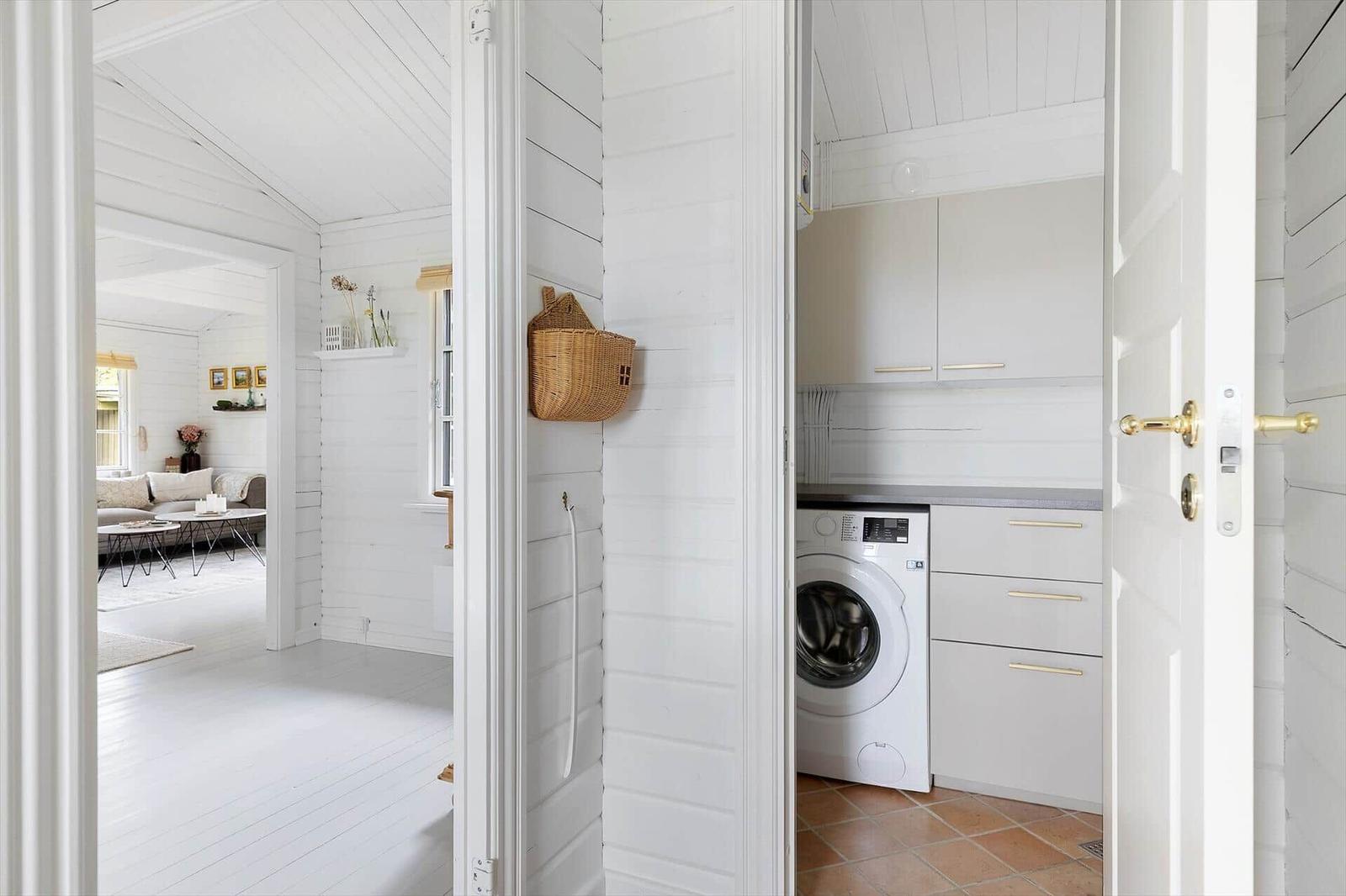 Laundry room with washing machine and cabinets. Corridor to living room with sofa and table.