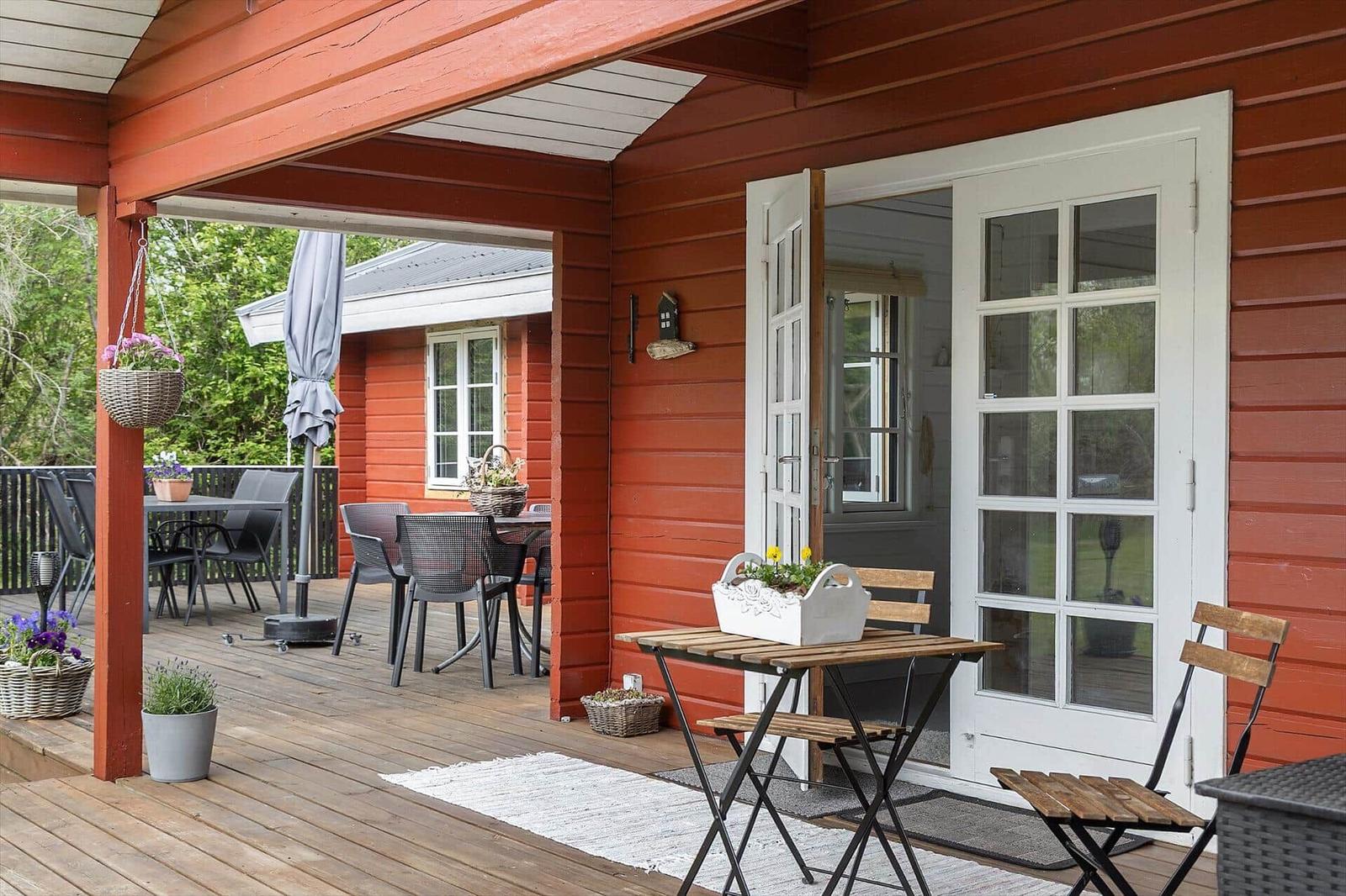 Red wooden house with terrace, tables, chairs, plants, and white doors.