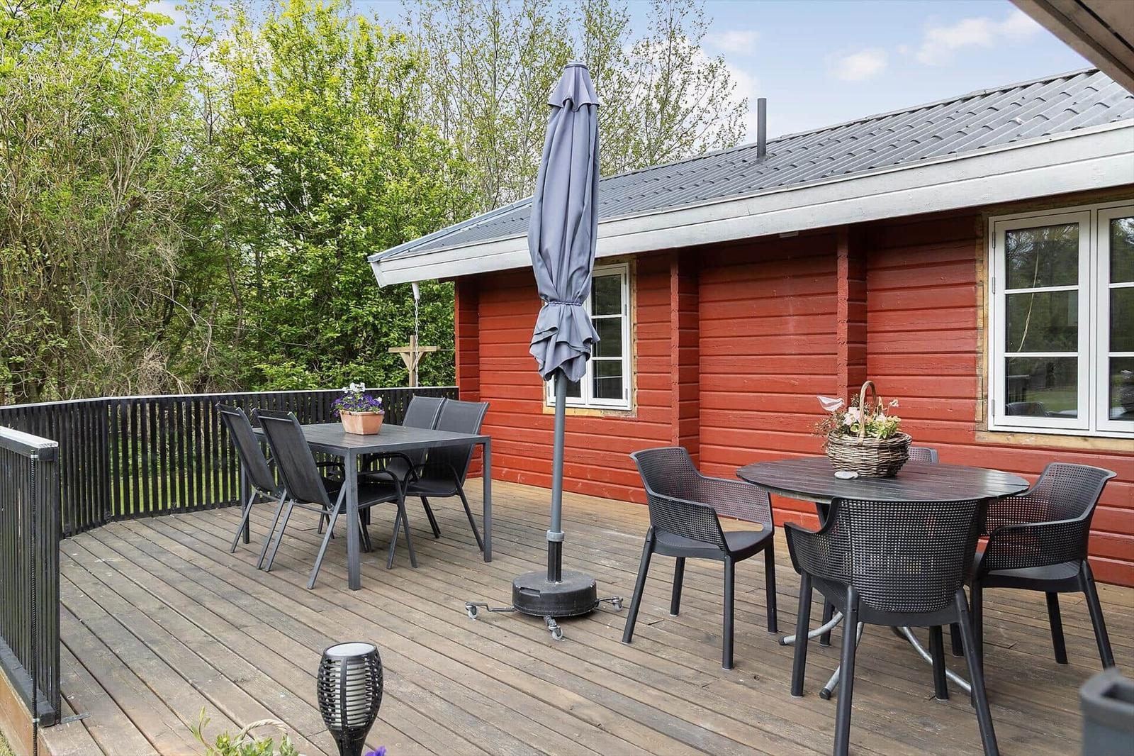 Deck with tables, chairs, and closed umbrella beside red wooden house.