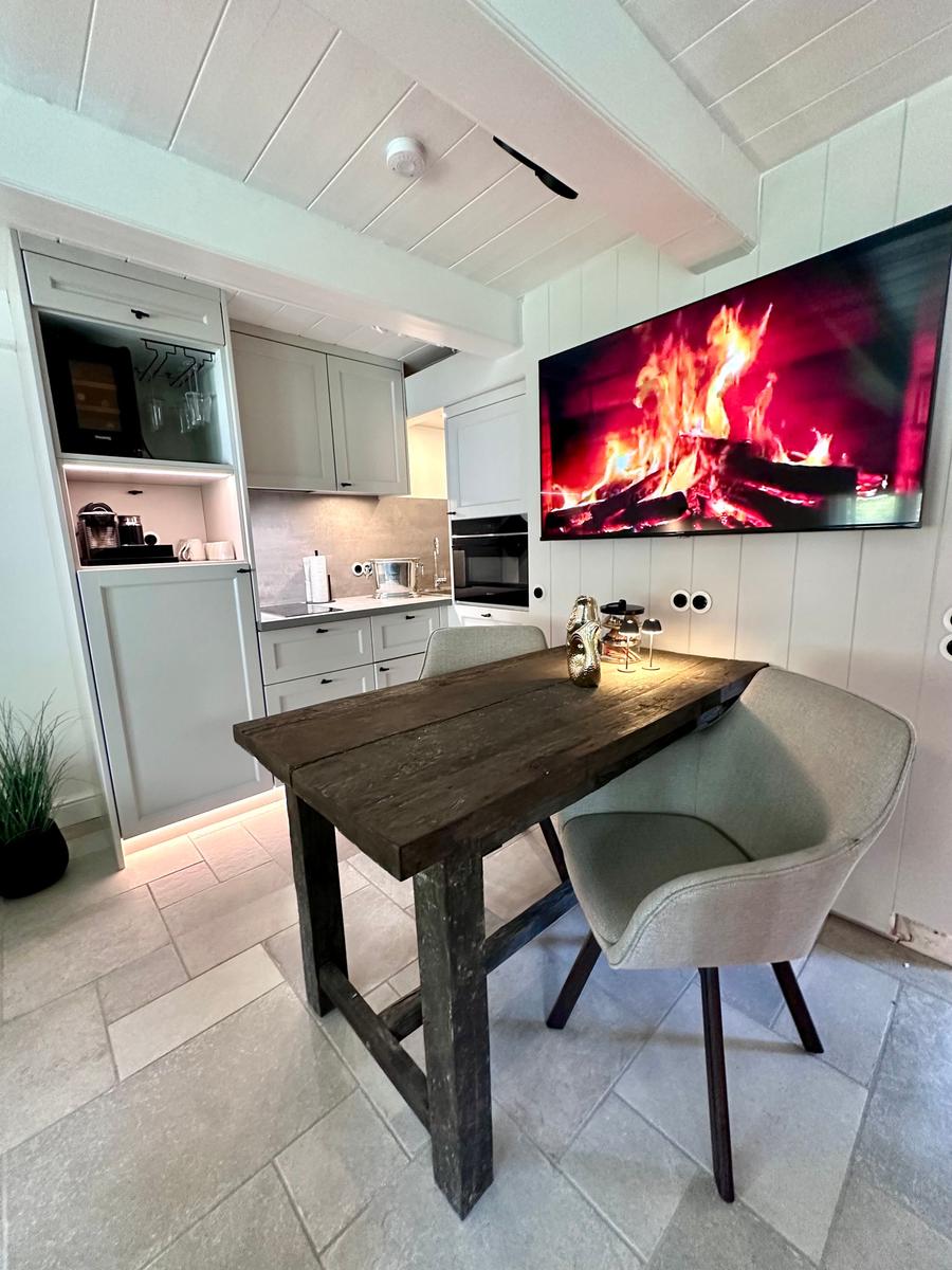 Essbereich mit Holztisch, zwei Stühlen und Küchenbereich. Wandfernseher zeigt Flammen.