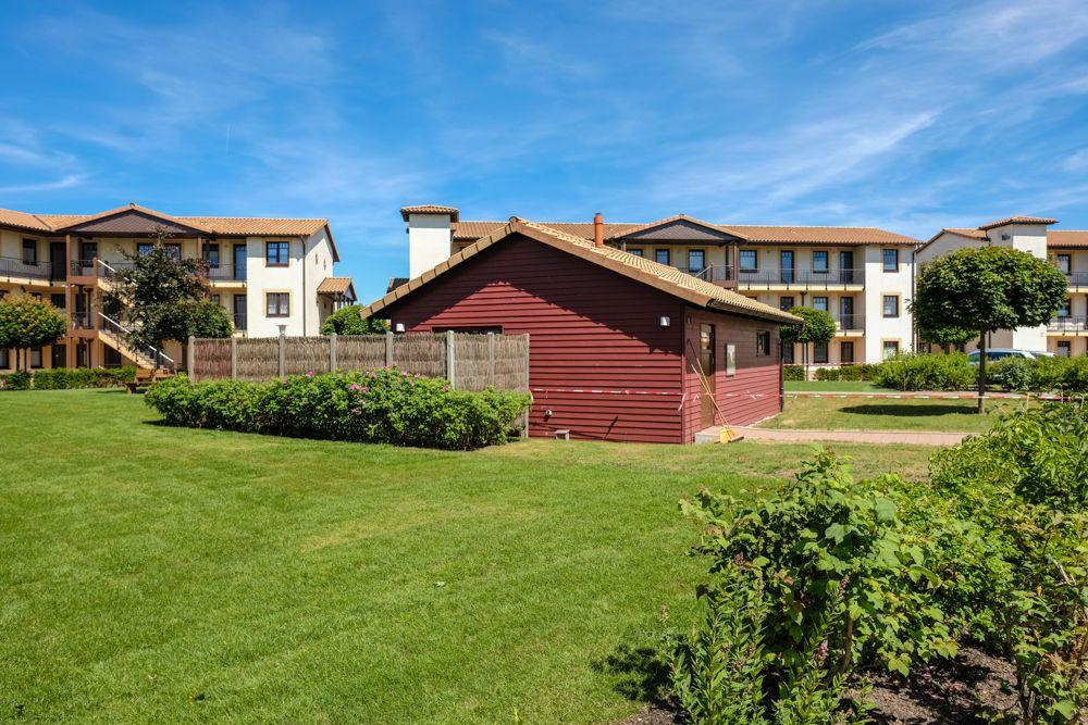 Roter Holzschuppen vor mehrstöckigem Wohnhaus mit Garten und Bäumen unter blauem Himmel.