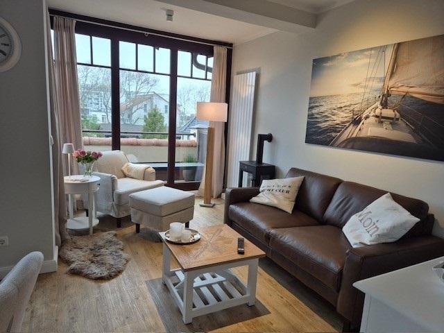 Wohnzimmer mit braunem Sofa, Holztisch und großem Fenster. Wandbild zeigt Segelyacht.