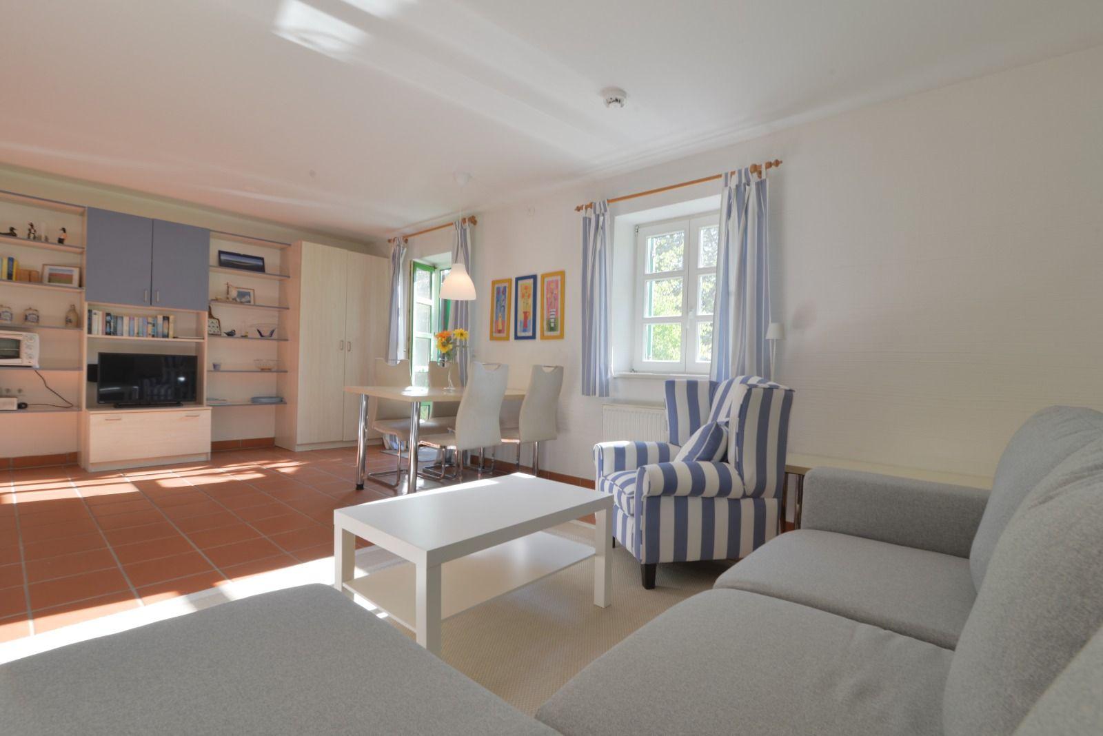 Wohn- und Essbereich mit Sofa, Stuhl und Tisch. Fenster mit Vorhängen. Wandregale und TV.