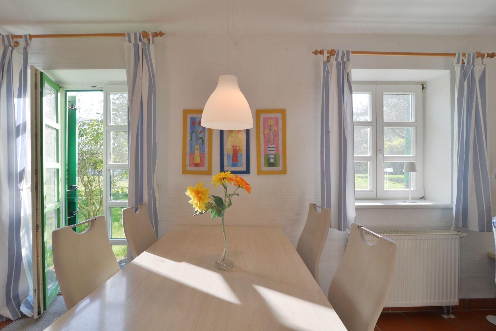 Esszimmer mit Tisch, Stühlen und Blumen. Fenster und Tür führen nach draußen.