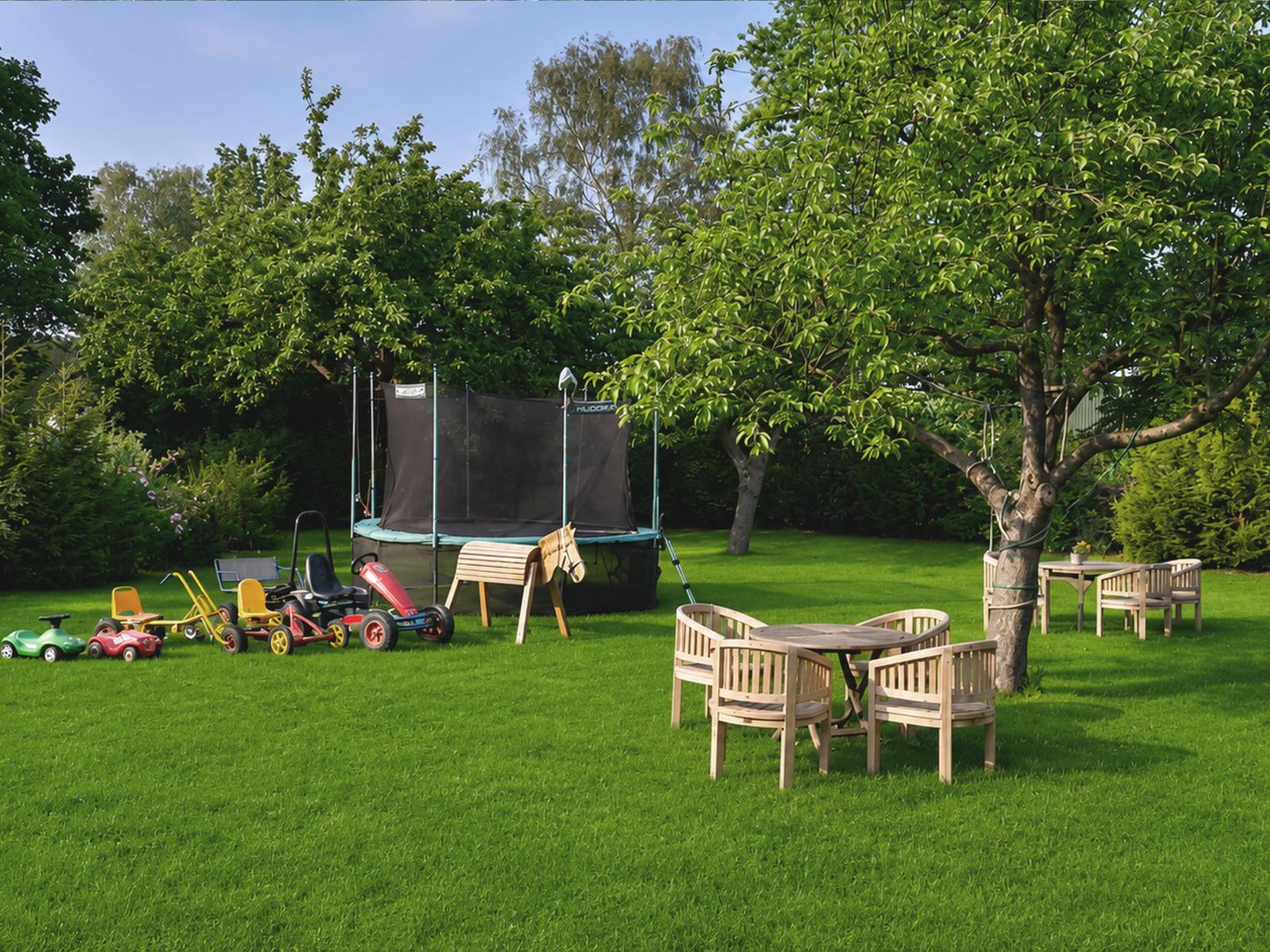 Garten mit Trampolin, Kinderspielzeug und Holztischen unter Bäumen.