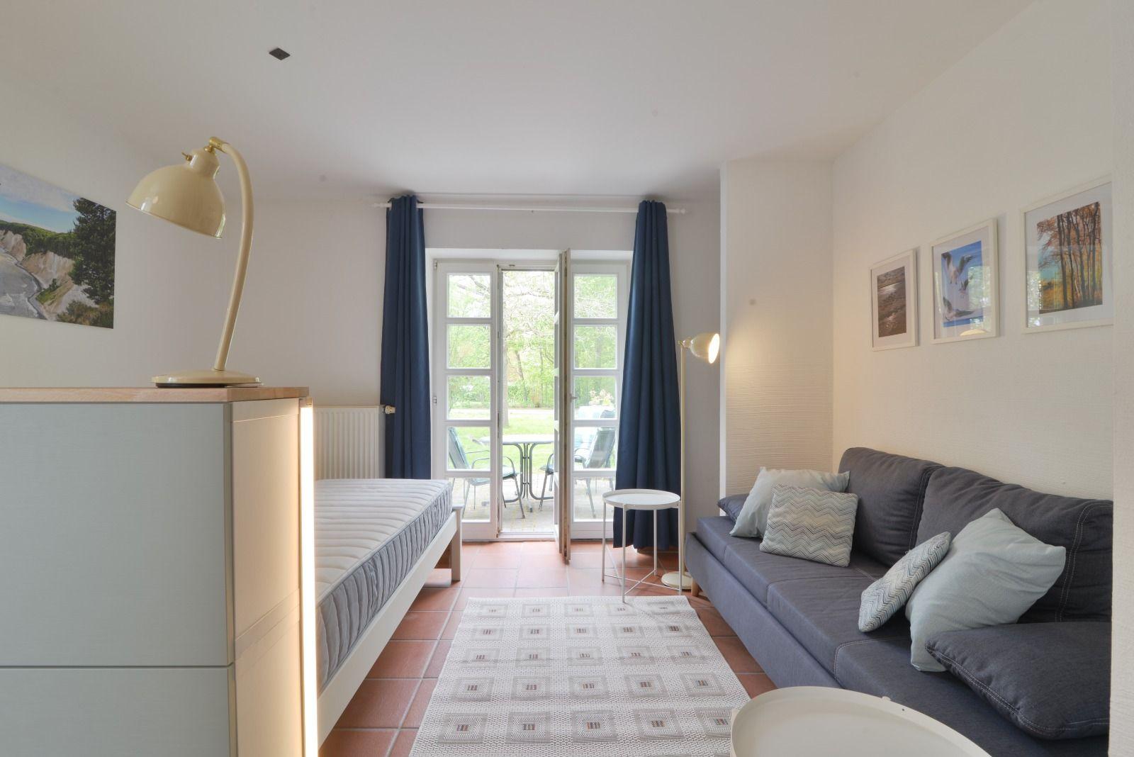 Ein Raum mit Sofa, Bett und Terrassentür. Wandbilder und Stehlampe sind sichtbar.