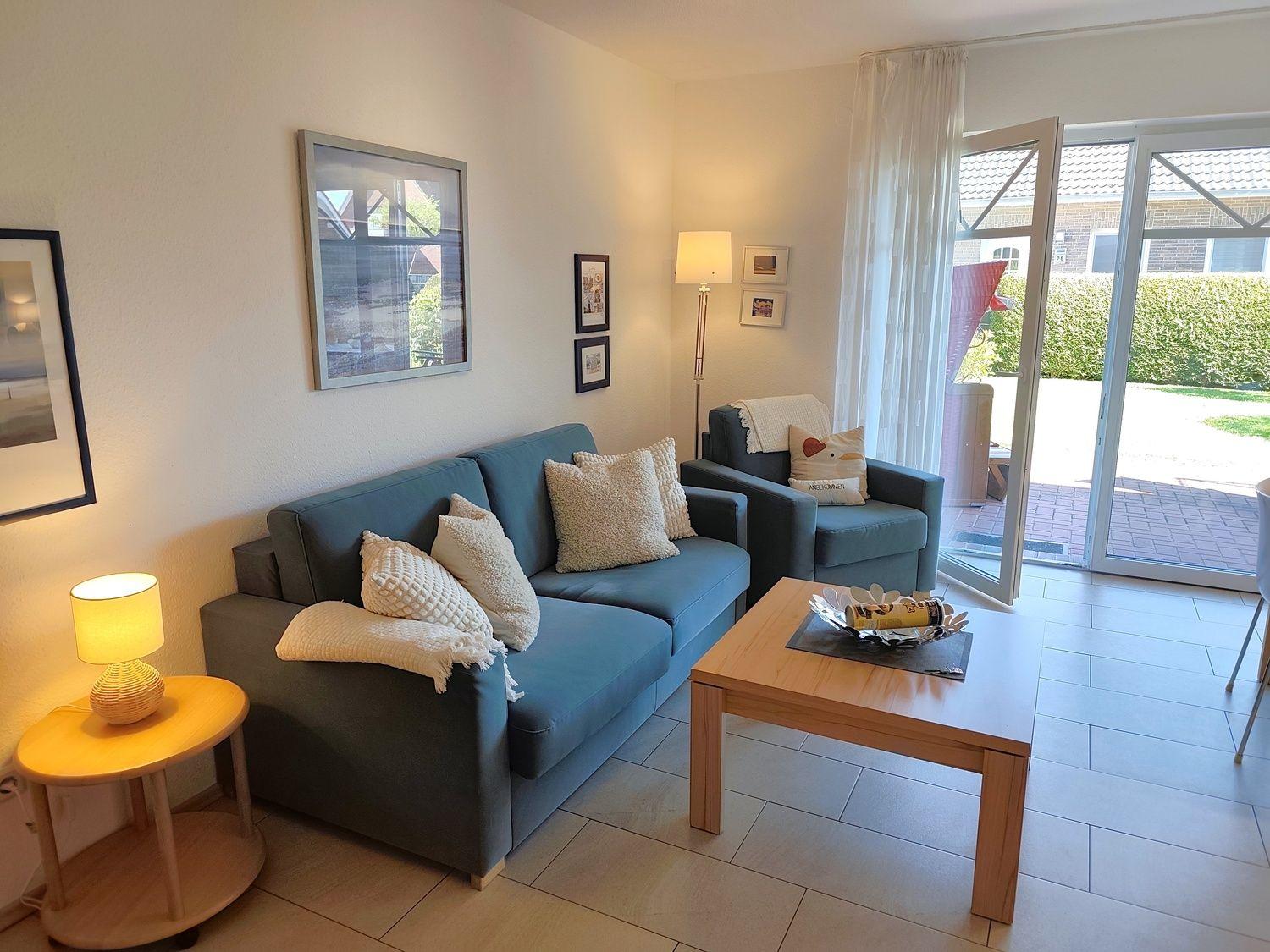 Wohnzimmer mit Sofa, Sessel und Tisch. Durchgang zur Terrasse mit Garten.