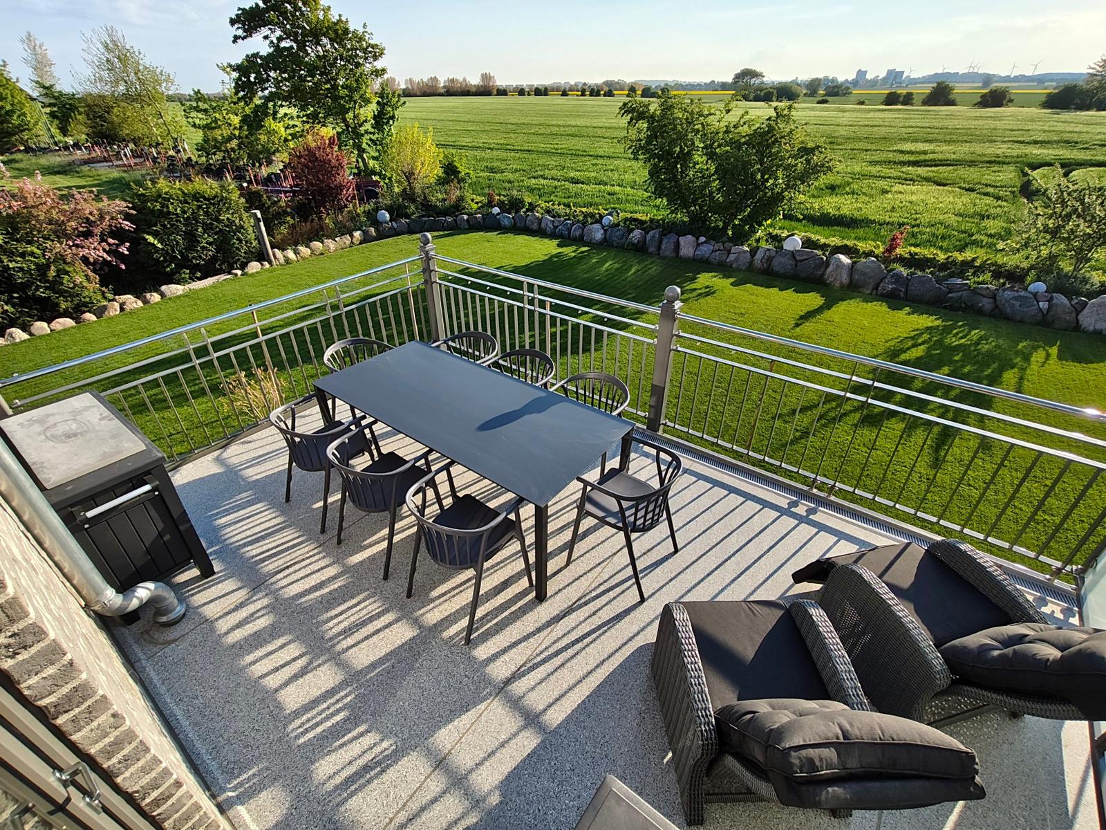 Terrasse mit Tisch, Stühlen und Liegen. Blick auf grüne Wiesen und Bäume.