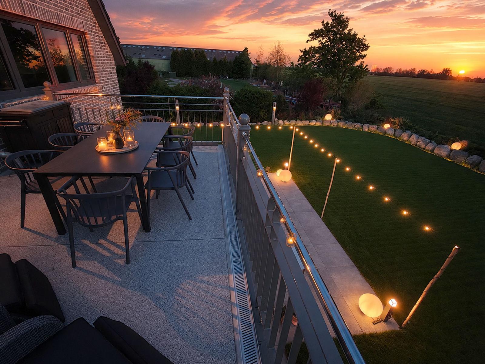 Terrasse mit Tisch, Stühlen und Lichterkette bei Sonnenuntergang. Grünfläche und Garten im Hintergrund.