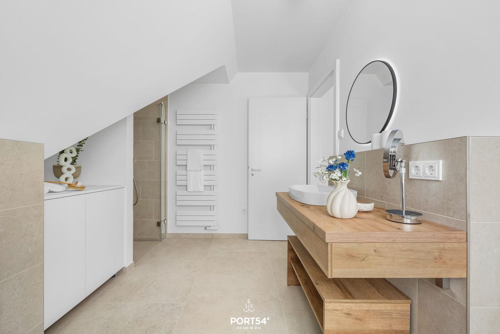 Bathroom with wooden vanity, round mirror, and shower under sloped ceiling.