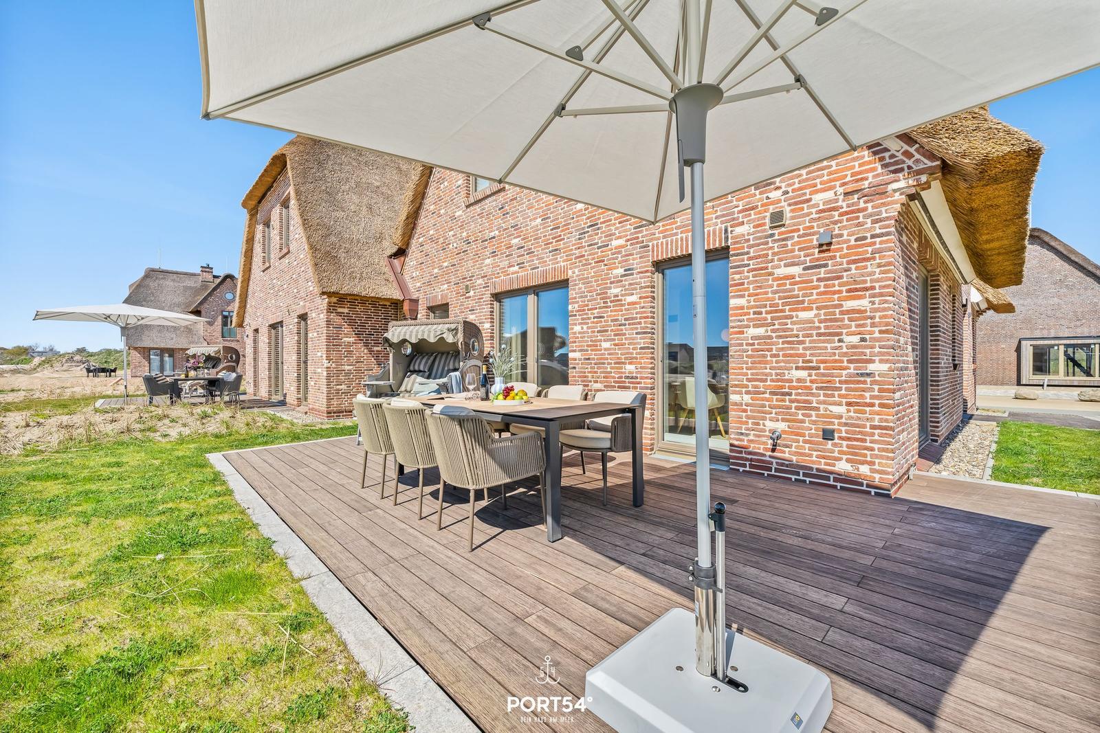 Deck with table, chairs, and umbrella next to a brick house with thatched roof.