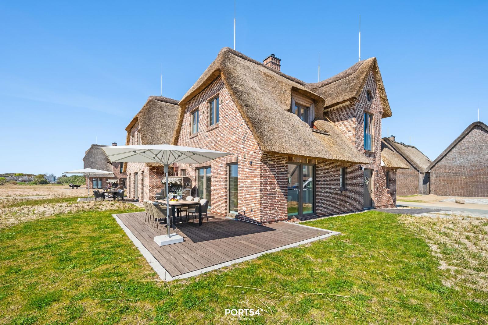 House with thatched roof and wooden terrace with table and umbrella.