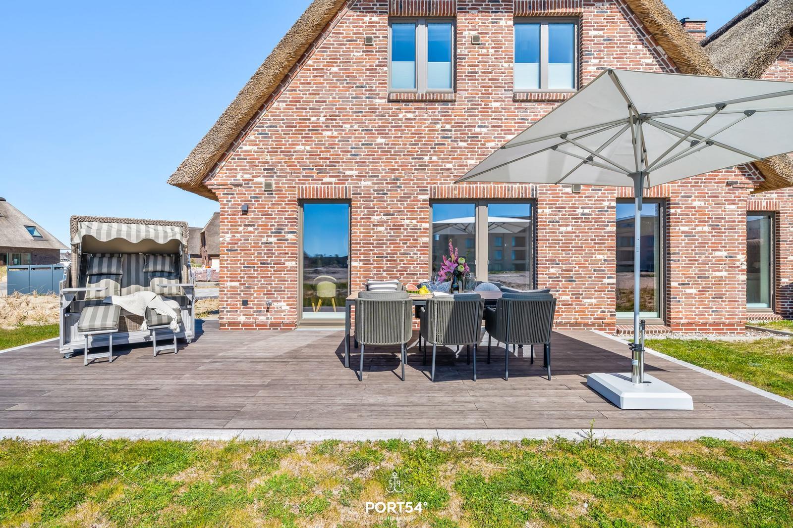 Terrasse mit Tisch, Stühlen und Sonnenschirm vor einem Backsteinhaus mit Strohdach.