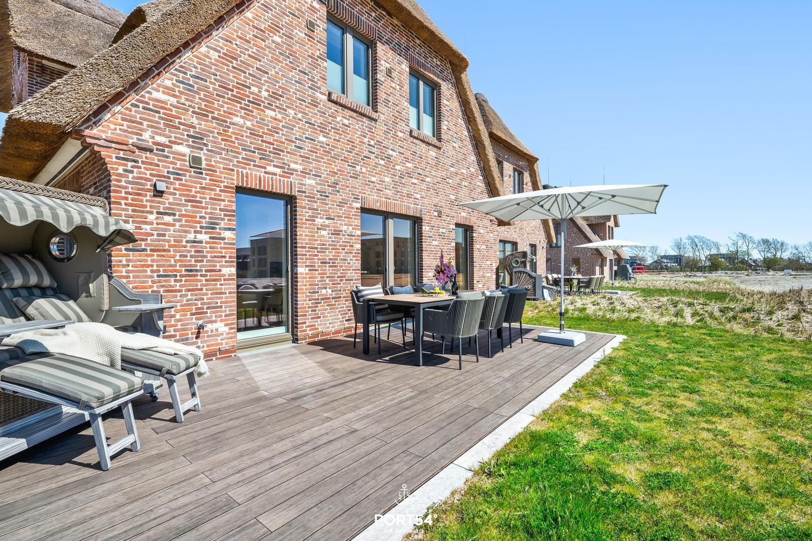 Terrasse mit Tisch, Stühlen und Sonnenschirm neben einem Backsteinhaus mit Strohdach.