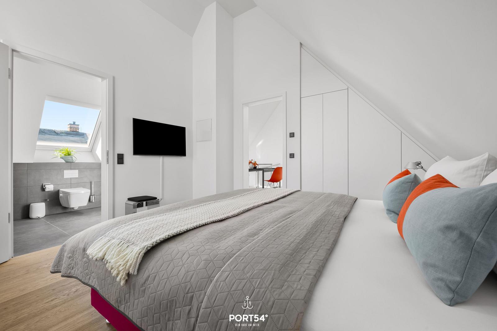 Schlafzimmer mit Dachschrägen, Bett, TV und angrenzender Toilette.