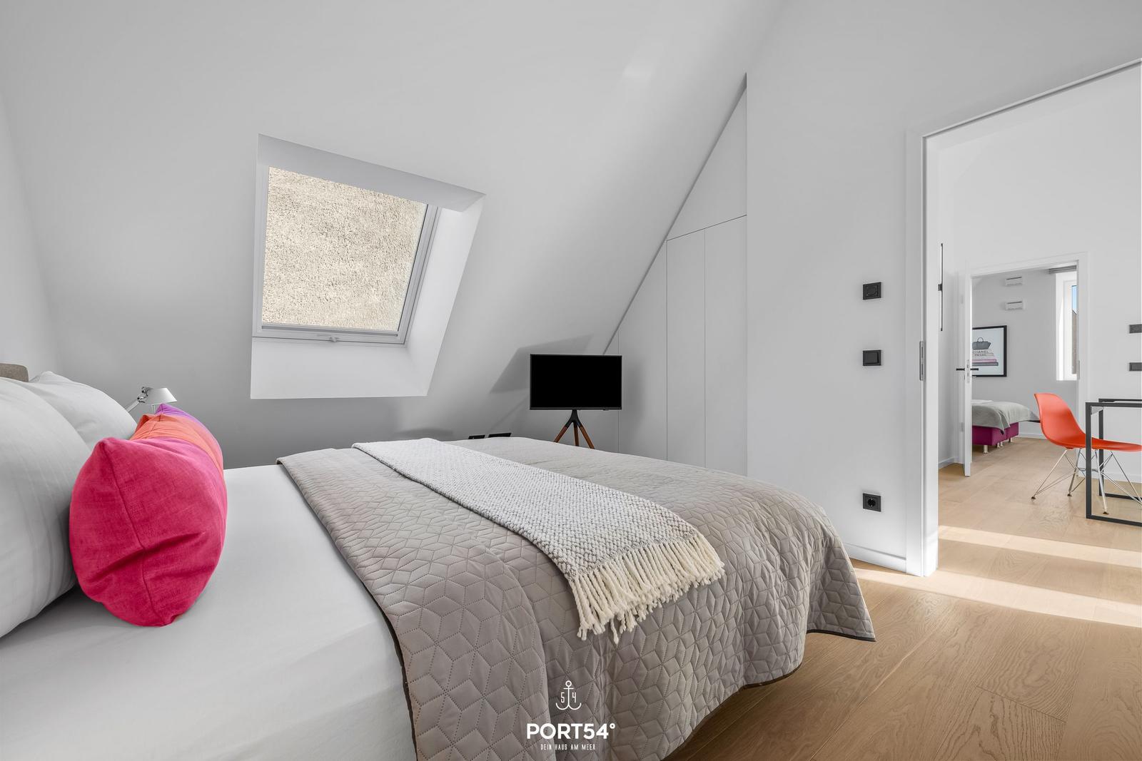 Schlafzimmer mit Dachschräge, Bett, Fenster und TV. Durchgang zu Wohnbereich mit Stuhl.