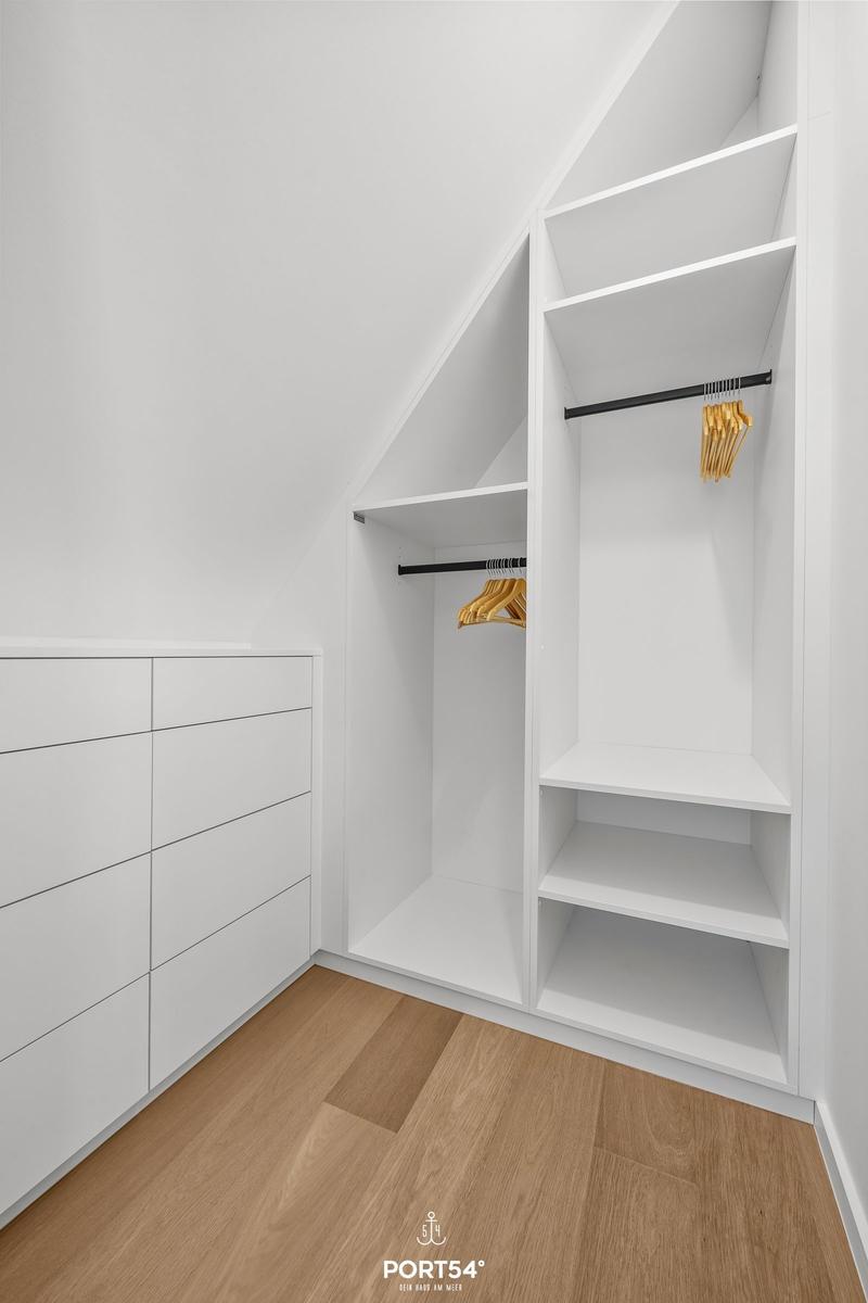 Weißer Schrank mit Kleiderstangen und Ablageflächen, Holzboden und schräge Decke.