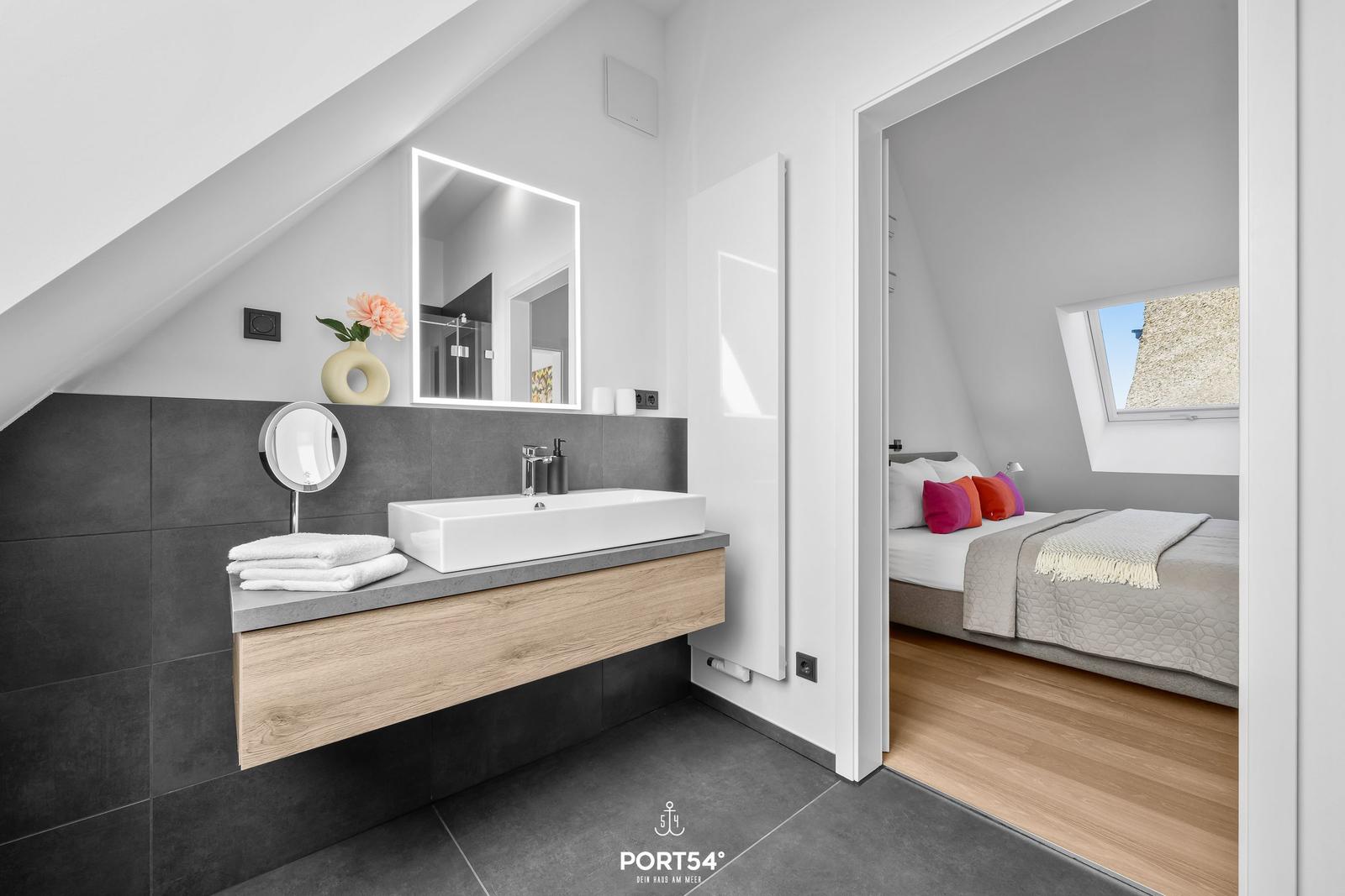 Badezimmer mit Waschtisch und Spiegel. Im Hintergrund ist ein Schlafzimmer mit Bett und Dachfenster.