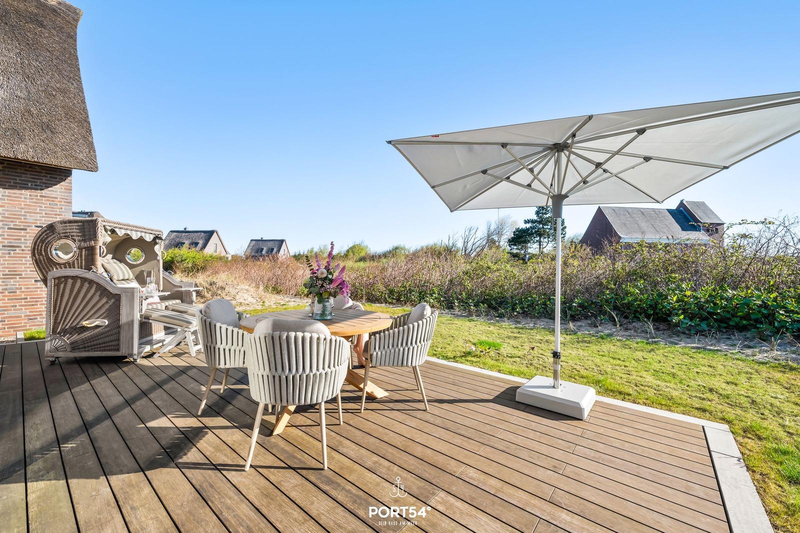 Terrasse mit Tisch, Stühlen und Sonnenschirm. Hintergrund: Gras, Sträucher und Häuser.