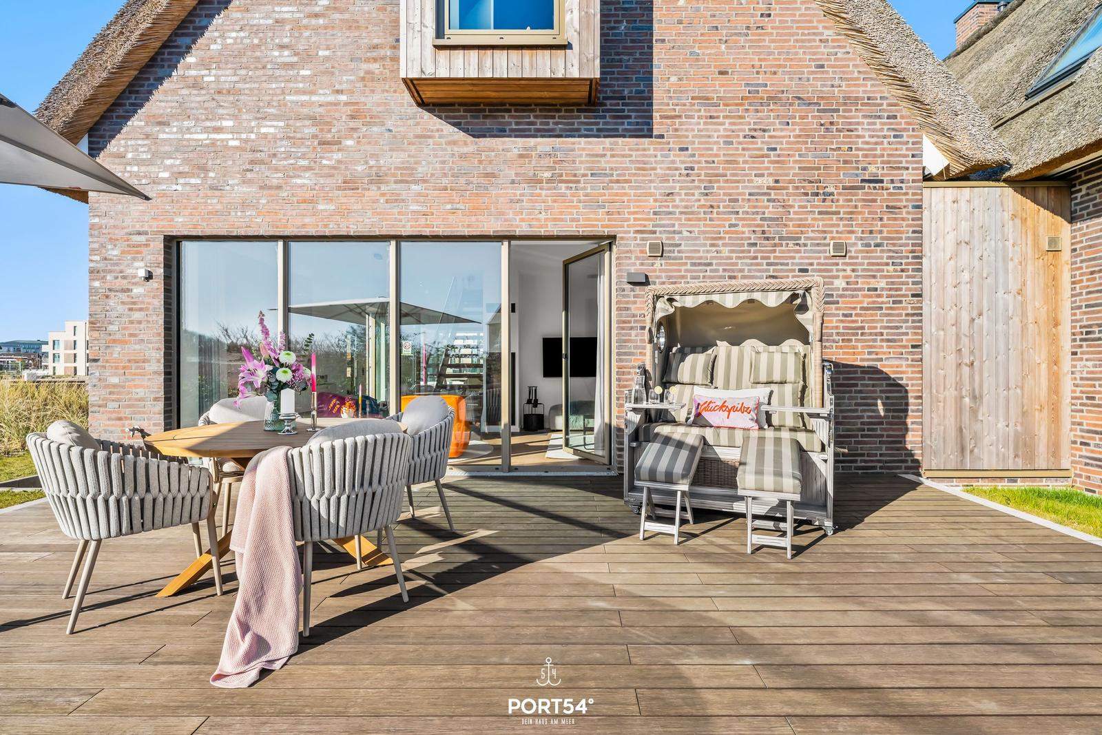 Terrasse mit Esstisch, Sitzgruppe und Liege vor Backsteinhaus mit Glasfront.