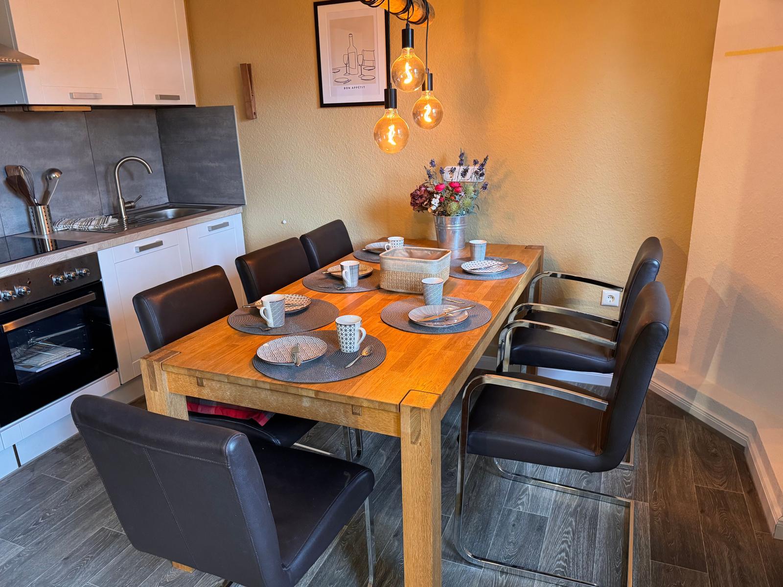 Essbereich mit Holztisch, sechs Stühlen und Küchenzeile im Hintergrund.