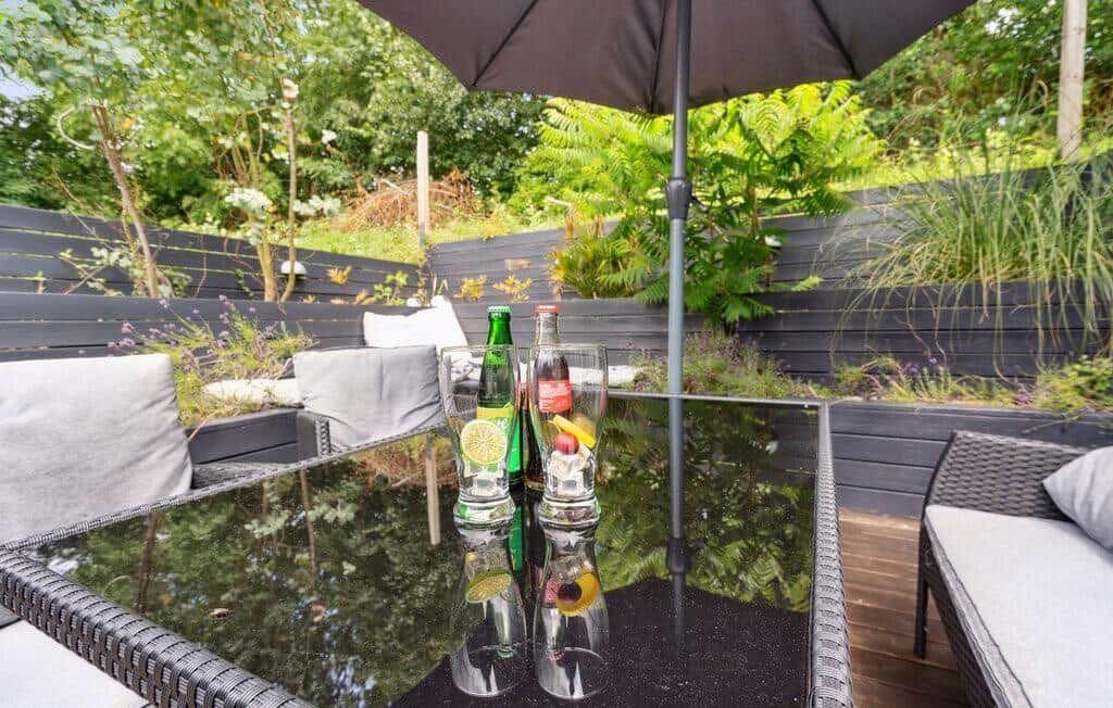 Terrace with table, seating, and umbrella. Background: green plants and wooden wall.