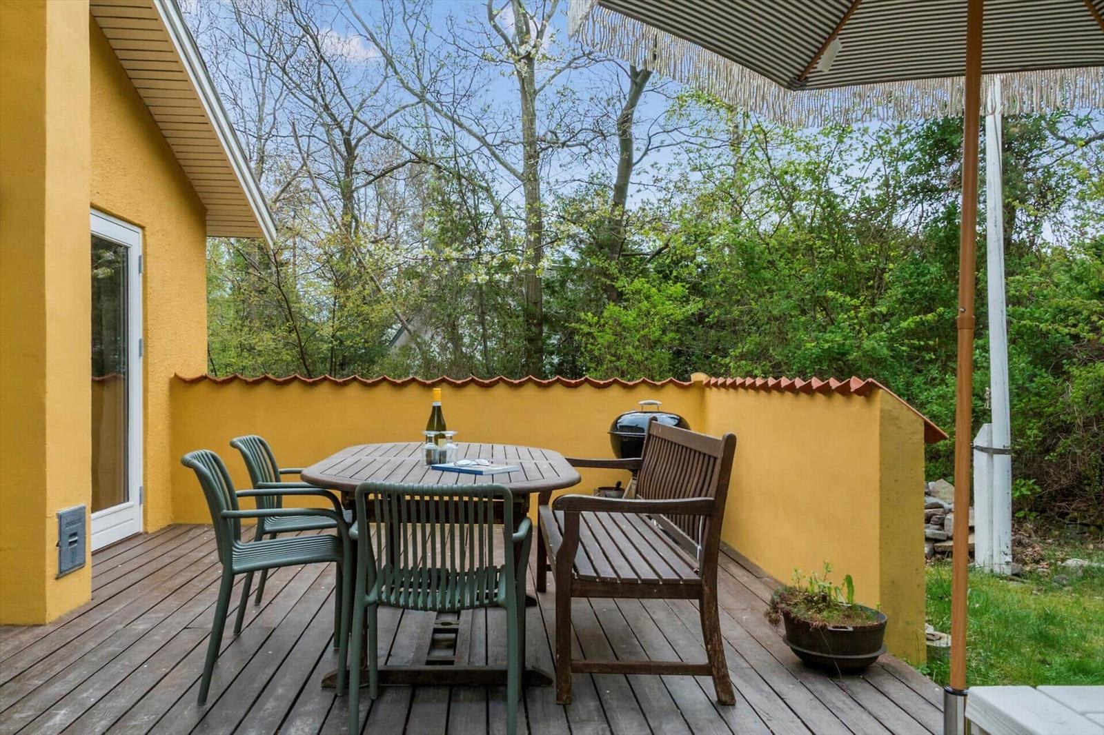 Terrace with table, chairs, and grill. Yellow stucco and wooden decking. View into green trees.