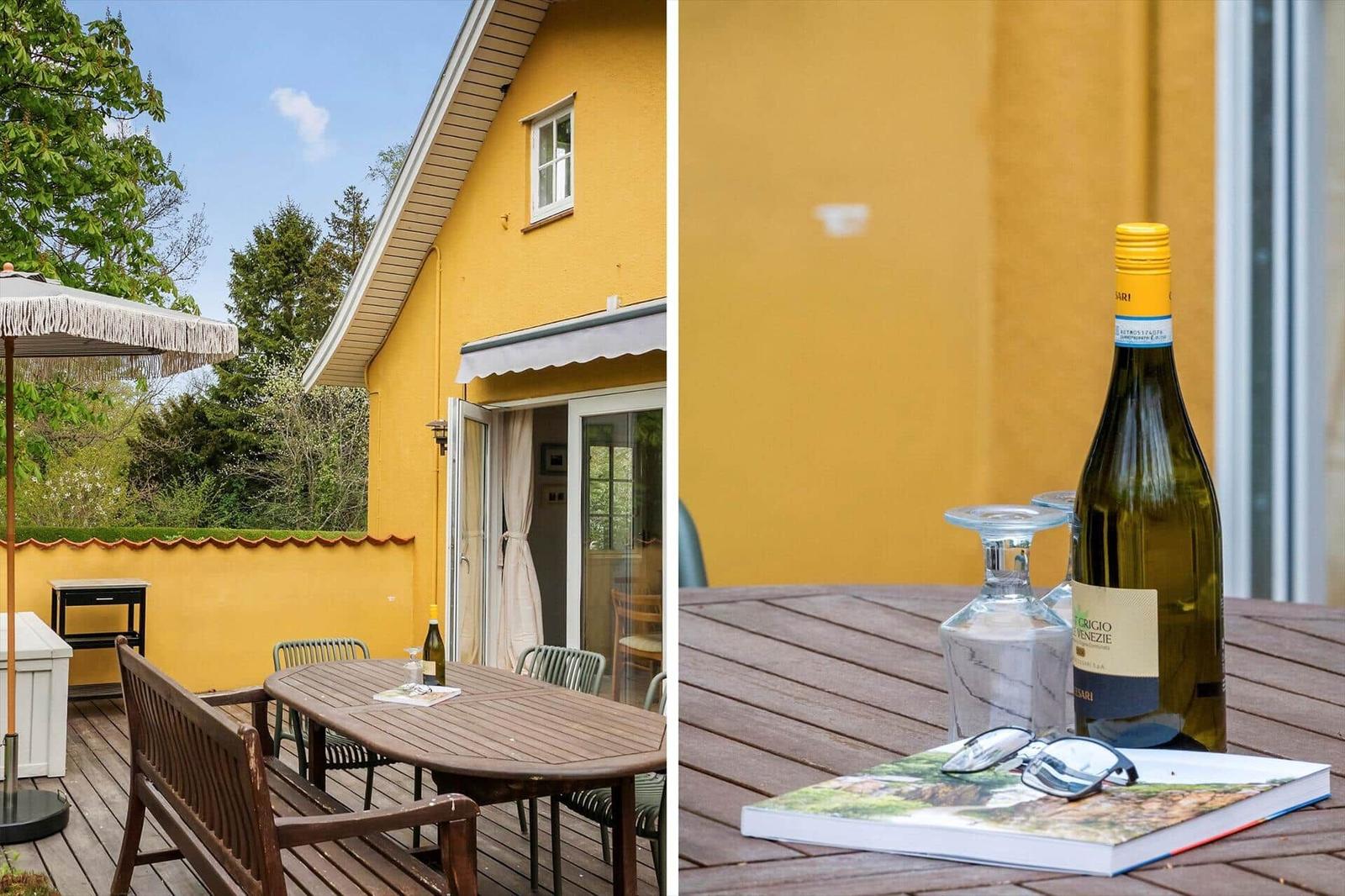 Terrace with table, chairs, and umbrella in front of yellow house. Table with wine bottle and book.