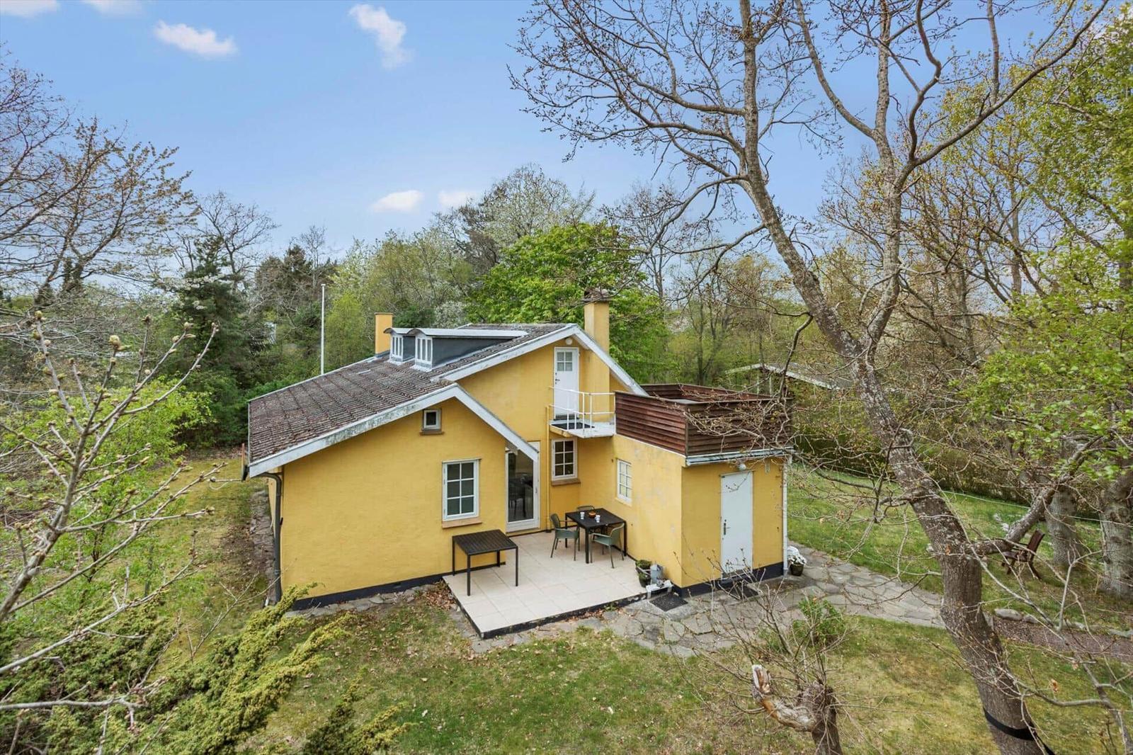 Yellow house with terrace and garden. Surrounded by trees under blue sky.