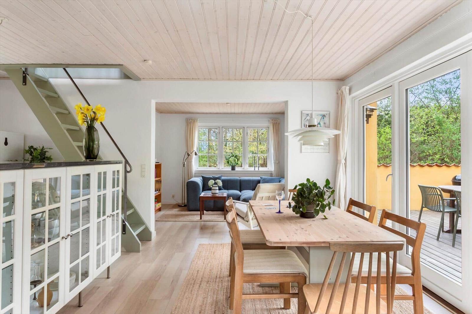 Dining area with wooden table and chairs, view into living room with sofa and windows.