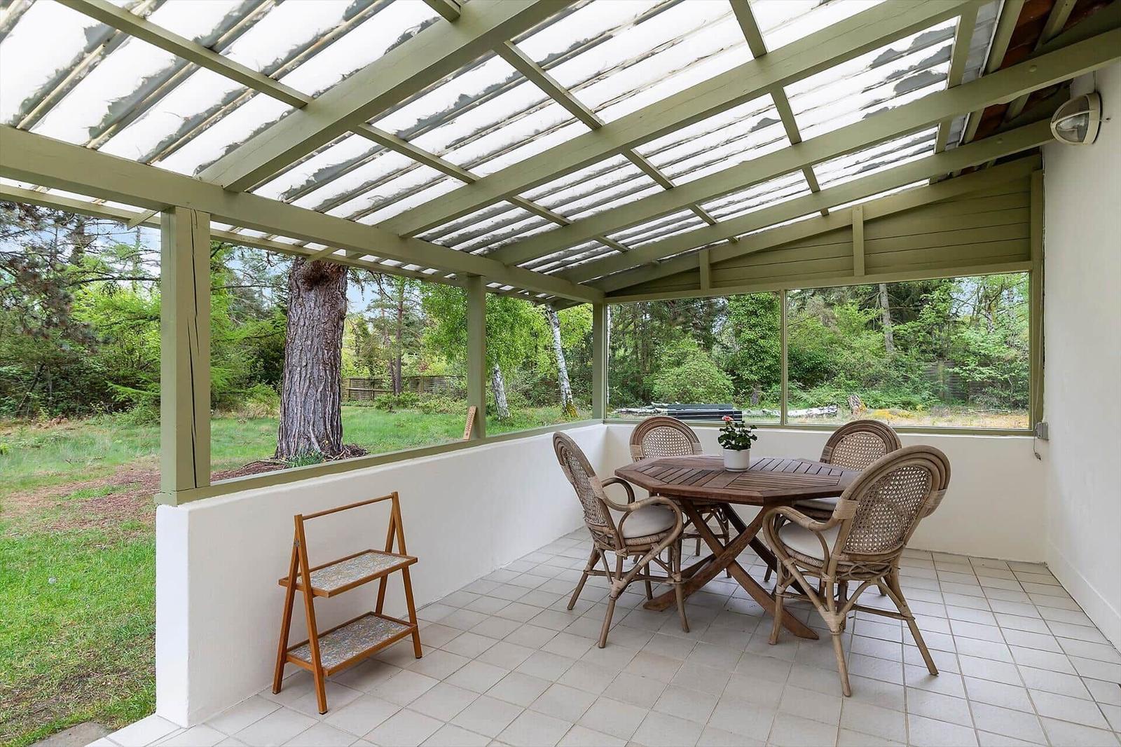 Patio with table and chairs under roof, view of green surroundings.