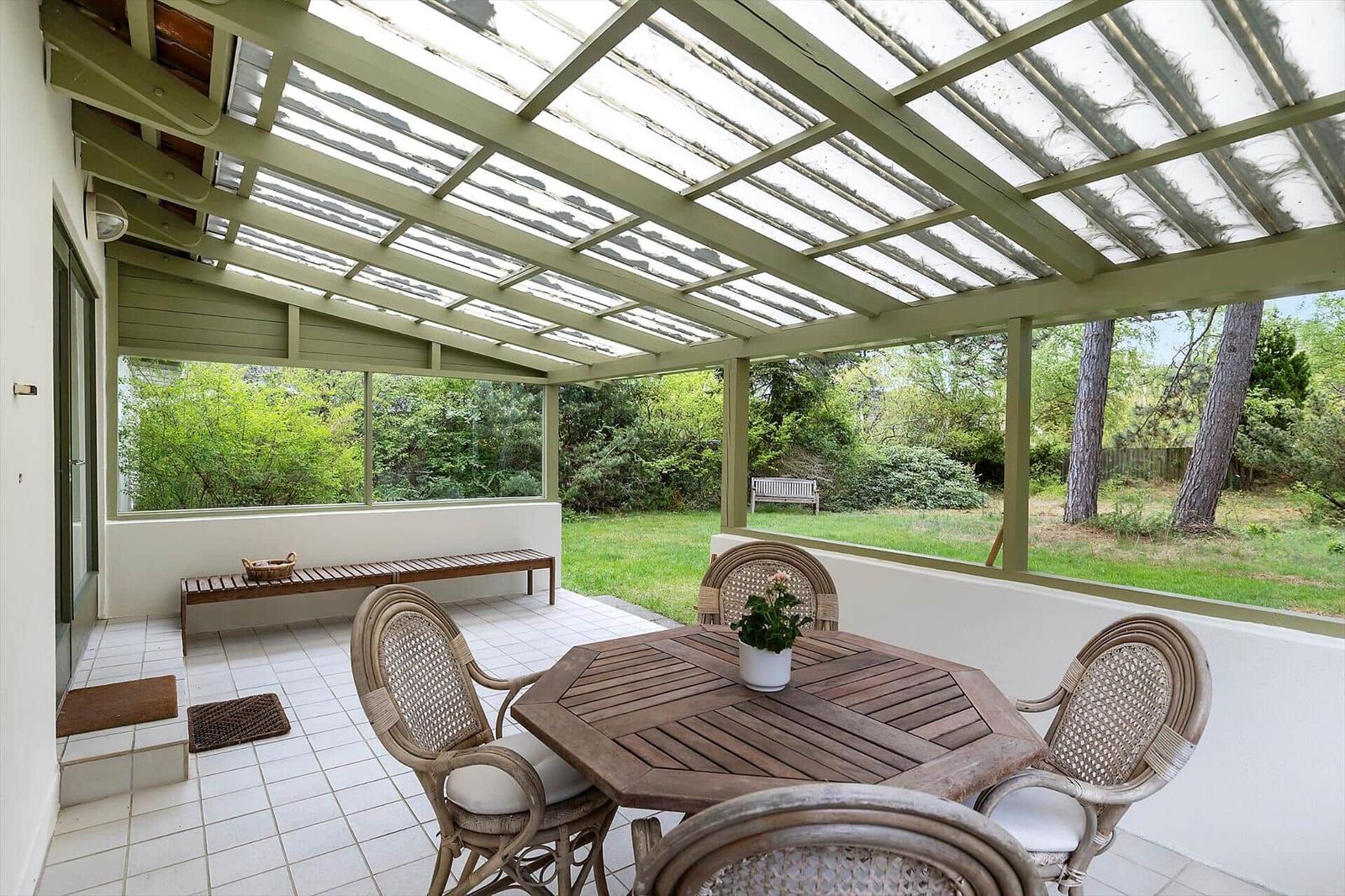 Terrace with table, chairs, and glass roof. View of green garden.
