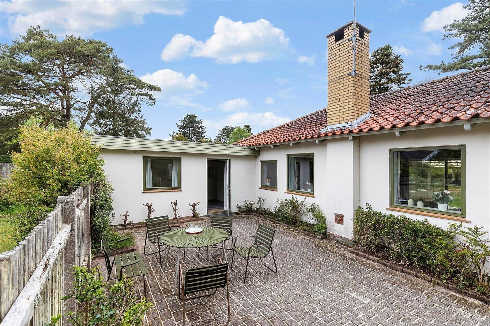 A house with white walls and red roof has a paved courtyard with table and chairs.