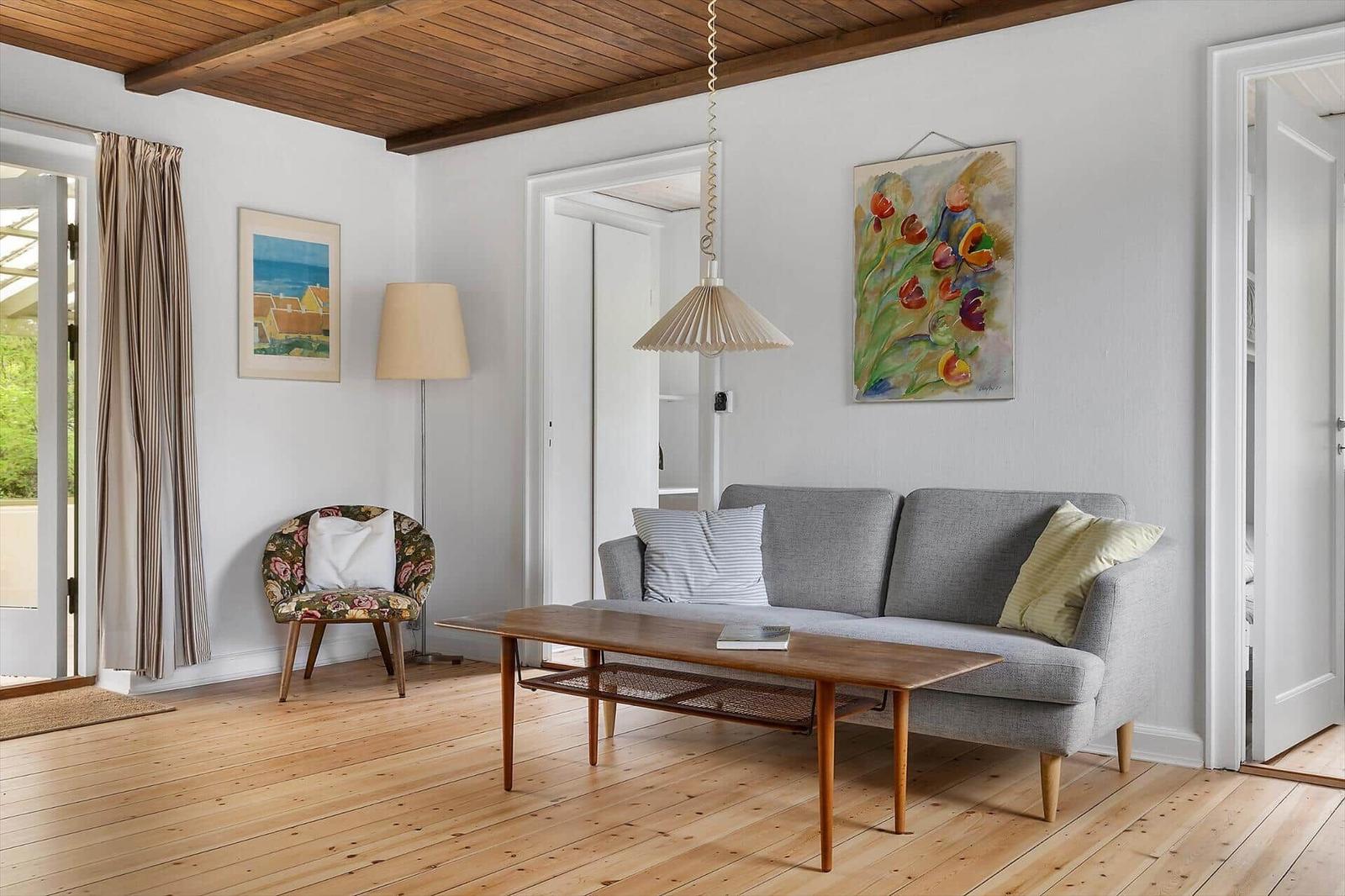 Living room with gray sofa, wooden floor, and ceiling beams. Two paintings hang on the wall.