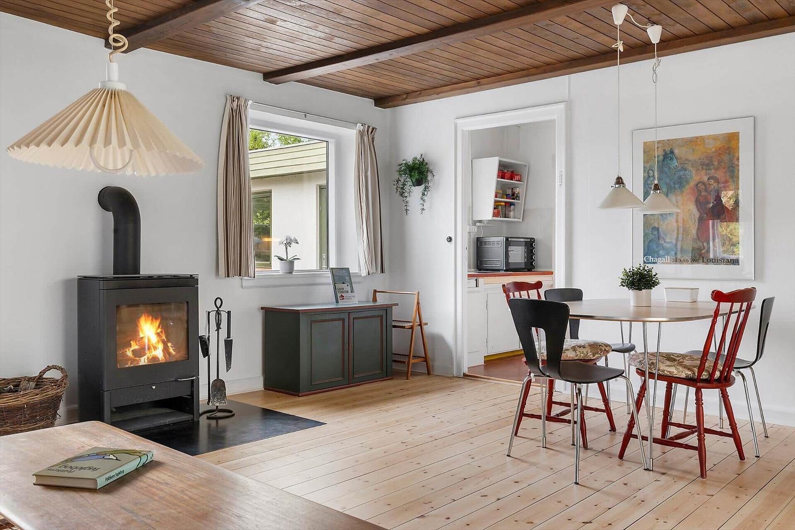 A room with fireplace, dining table, and kitchen area. Wooden ceiling and floor.