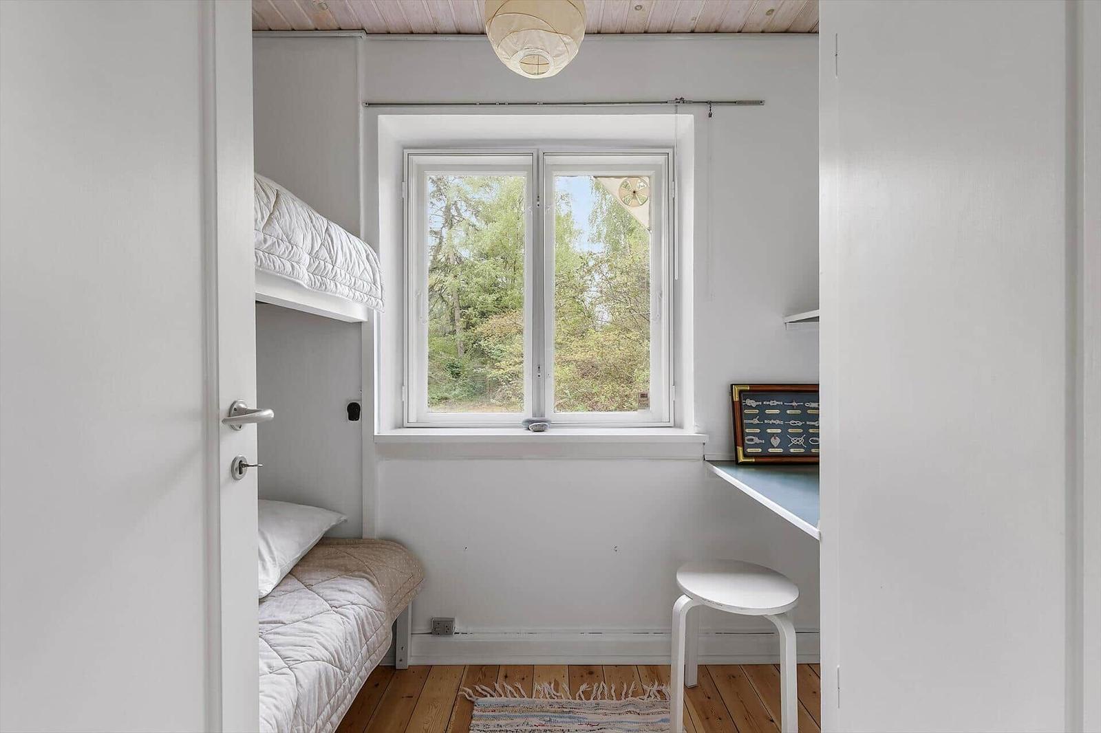 Room with bunk bed, window, and desk. Wooden floor and white paint.