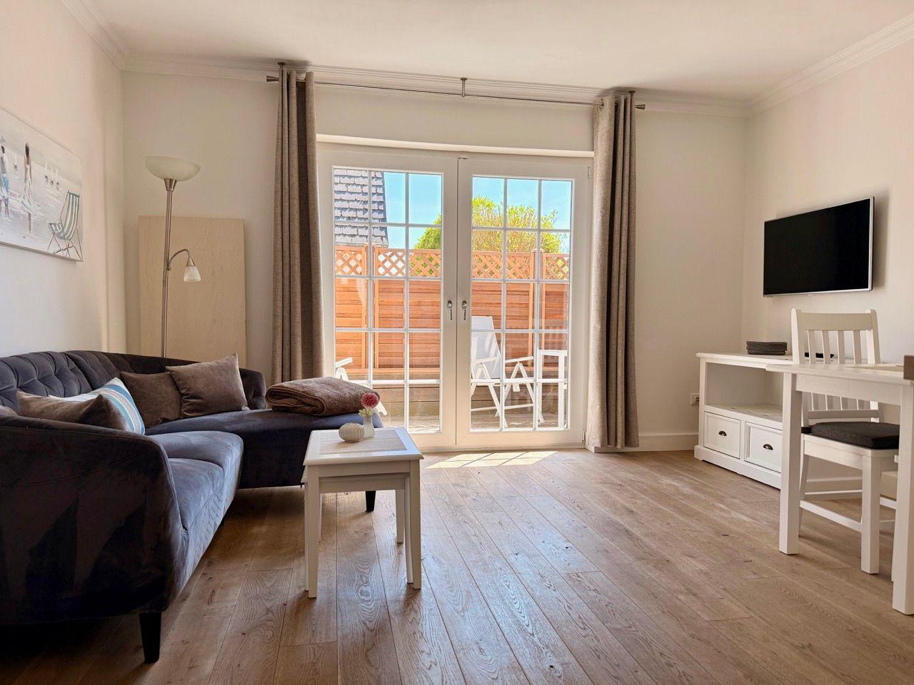 Wohnzimmer mit Sofa, Tisch und TV. Durchgehende Glastür zum Balkon mit Garten.