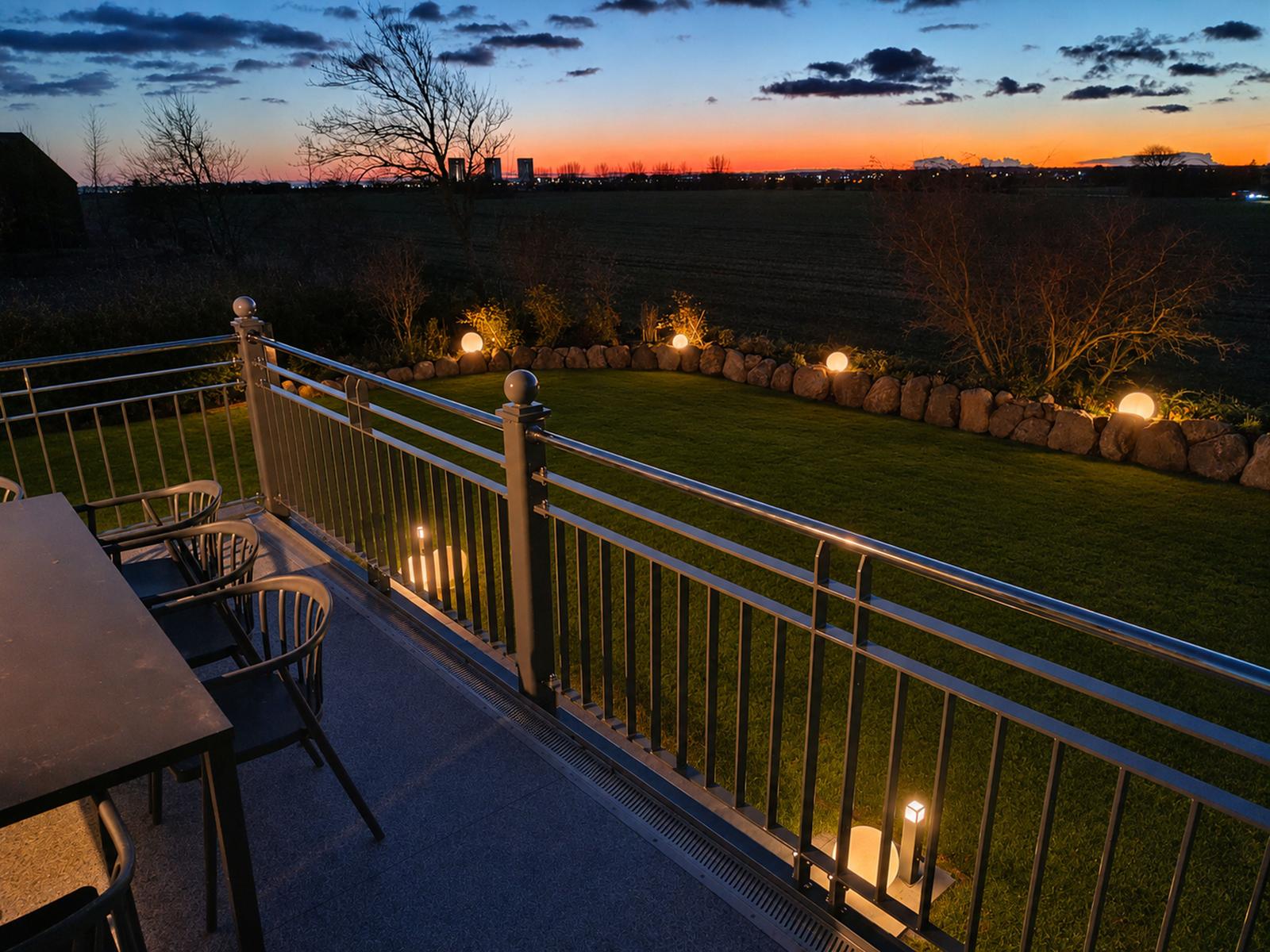 Terrasse mit Tisch und Stühlen, beleuchteter Garten und Landschaft bei Sonnenuntergang.