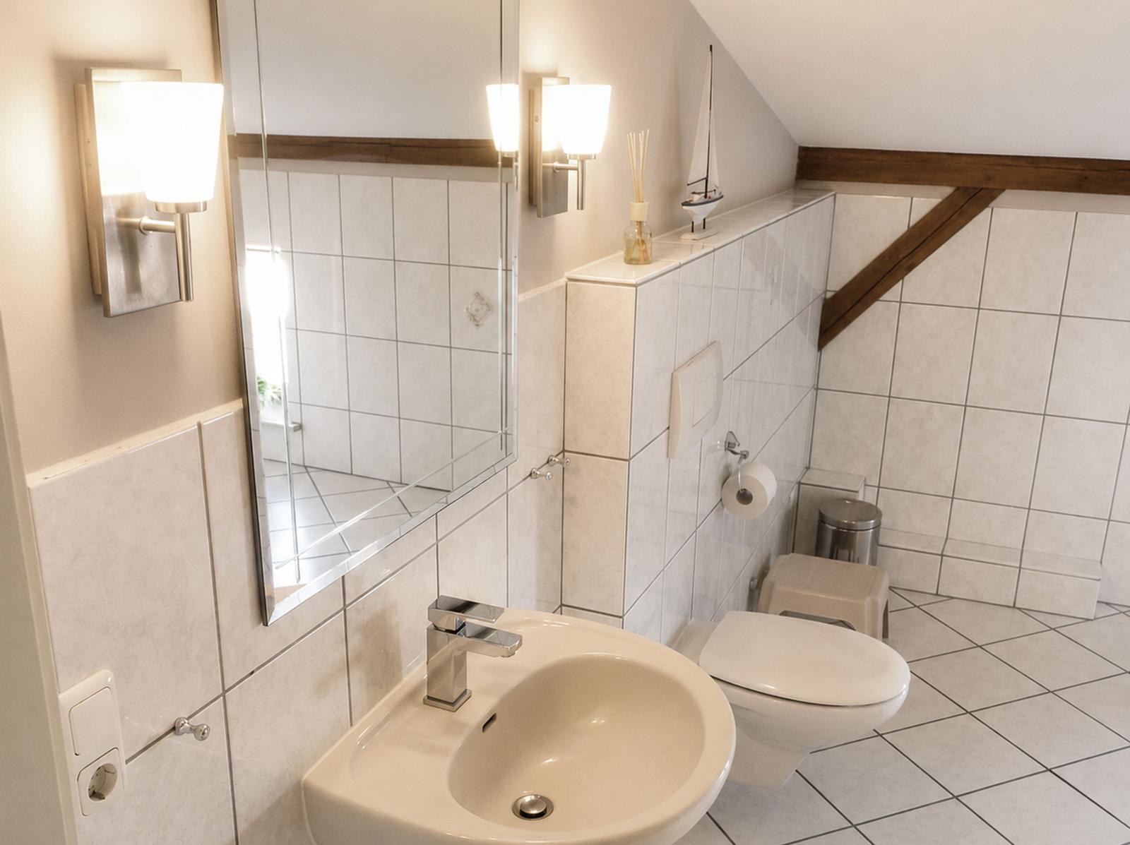 Badezimmer mit Waschbecken, Spiegel und Toilette unter schräger Decke mit Holzbalken.