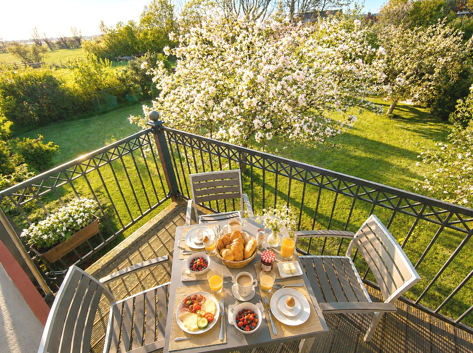 Frühstückstisch auf Balkon mit Blick auf grünen Garten und blühenden Baum.