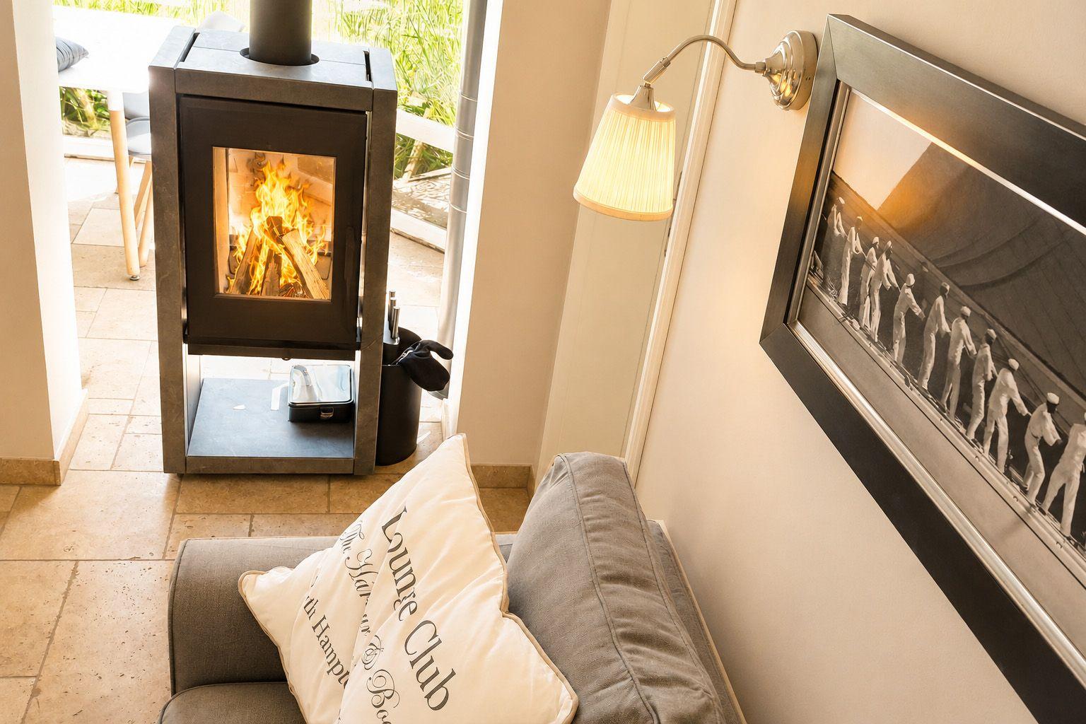 Ein Kaminofen mit brennendem Holz, ein Sofa mit Kissen und eine Wandlampe neben einem Bild.