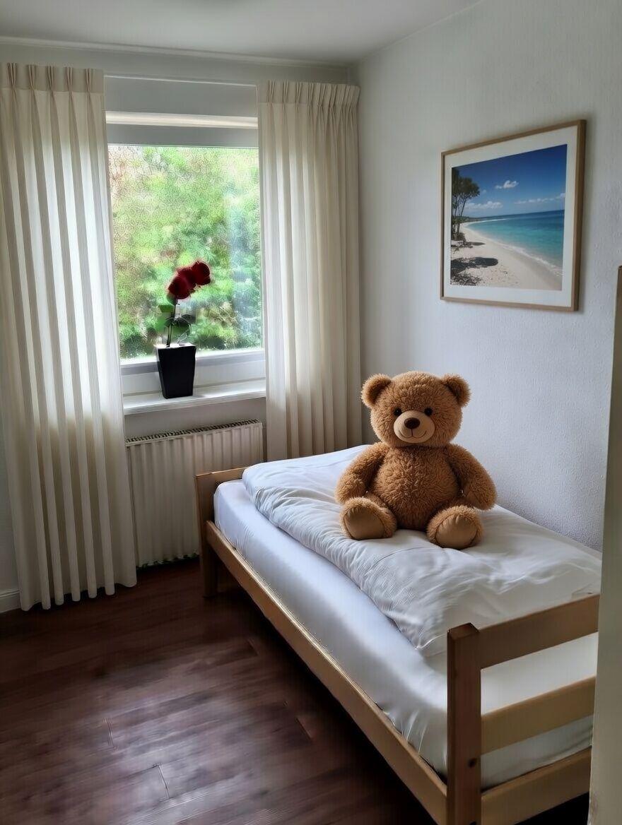 Ein Bett mit einem Plüschbären darauf, ein Fenster mit Blick auf Grün und eine Wanddekoration.