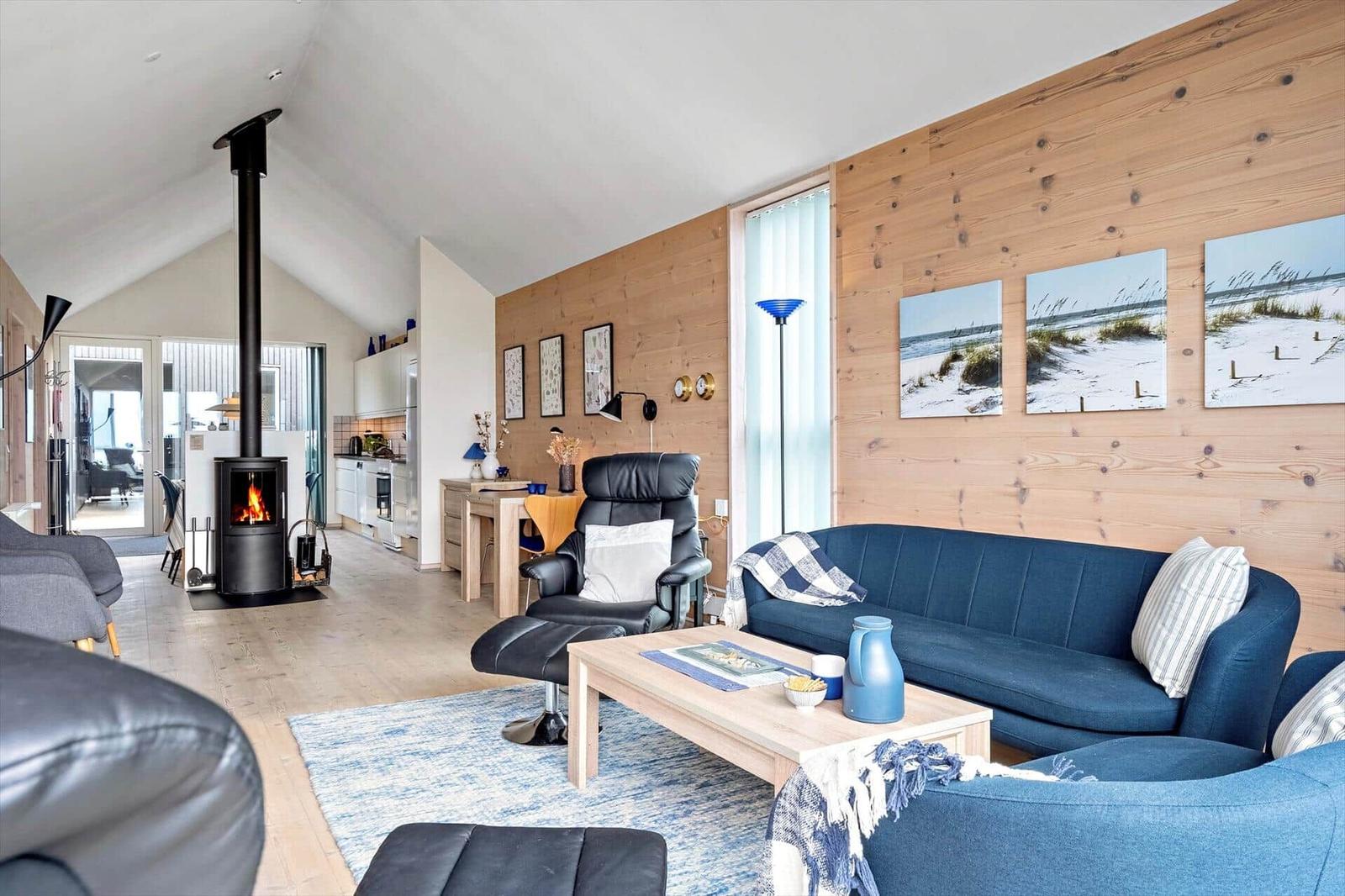 Wohnzimmer mit Kamin, Sofa und Küchenbereich. Holzwand und Strandbilder.