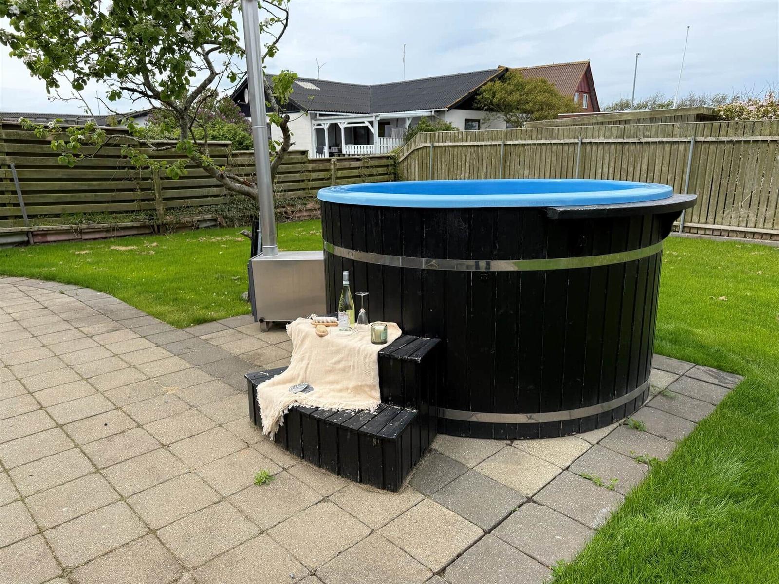 Schwarzer Holzhot tub mit blauem Rand auf gepflastertem Hof, umgeben von Gras und Holzzaun.